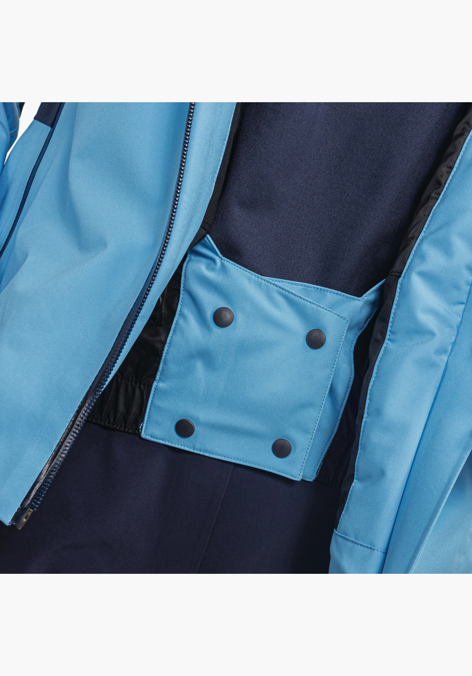Jacket Style Trifex WMS montre la jupe pare-neige intérieure bleue avec quatre boutons-pression foncés et une bande élastique à côté de la fermeture éclair frontale, gros plan sur fond blanc/neutre.