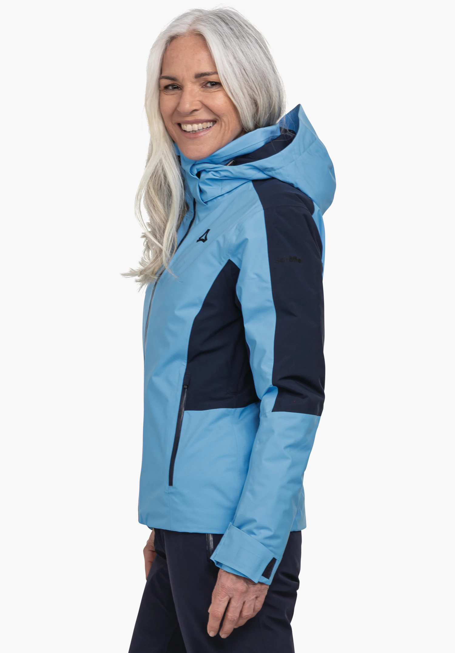 Jacket Style Trifex WMS veste outdoor: femme aux cheveux blanc clair porte une veste blanc clair et noire, sourit de profil devant fond blanc. Texte sur la manche: "Schöffel".