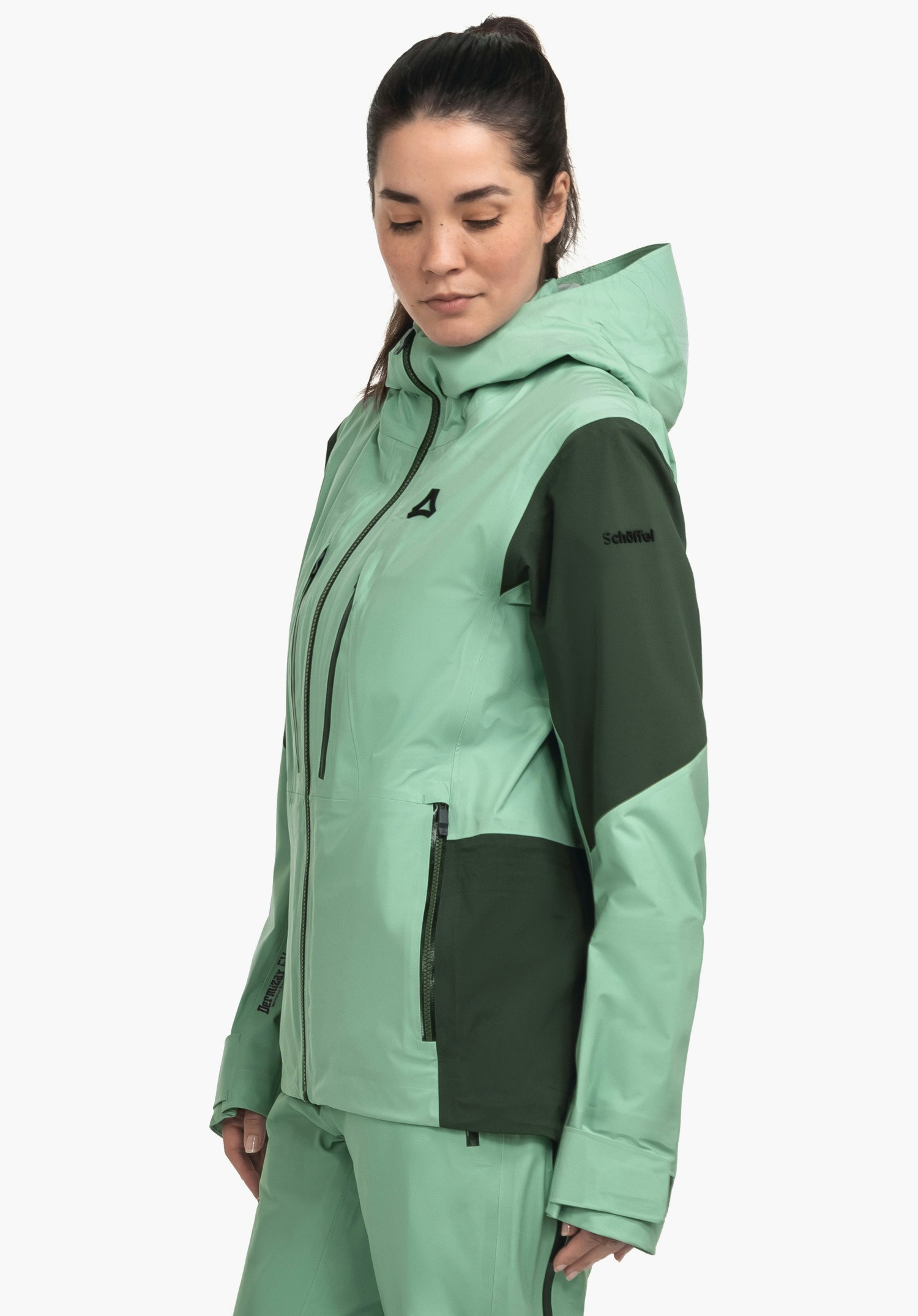 Shell Jk Style Lufeld WMS Jacke (grün hell mit grün dunkel Akzenten), getragen von einer Frau in seitlicher Pose, sie blickt nach unten, mit Kapuze vor weißem Studiohintergrund. Text: "Schöffel". Schöffel-Logo.