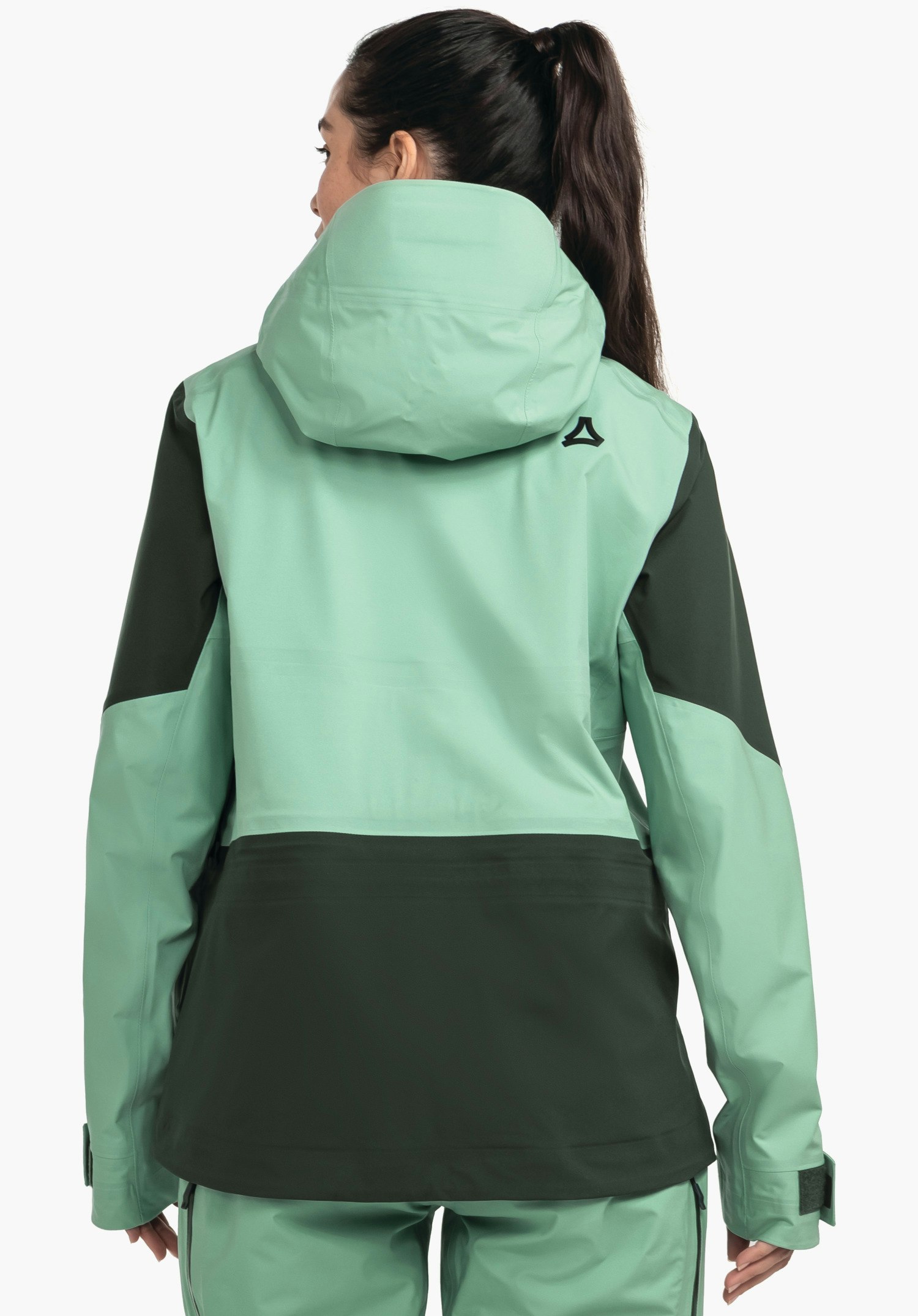 Shell Jk Style Lufeld WMS Jacke: Frau trägt hellgrüne und dunkelgrüne Regenjacke, steht mit Rücken zur Kamera, Kapuze aufrecht, Schöffel-Logo auf rechter Schulter, vor weißem Hintergrund.