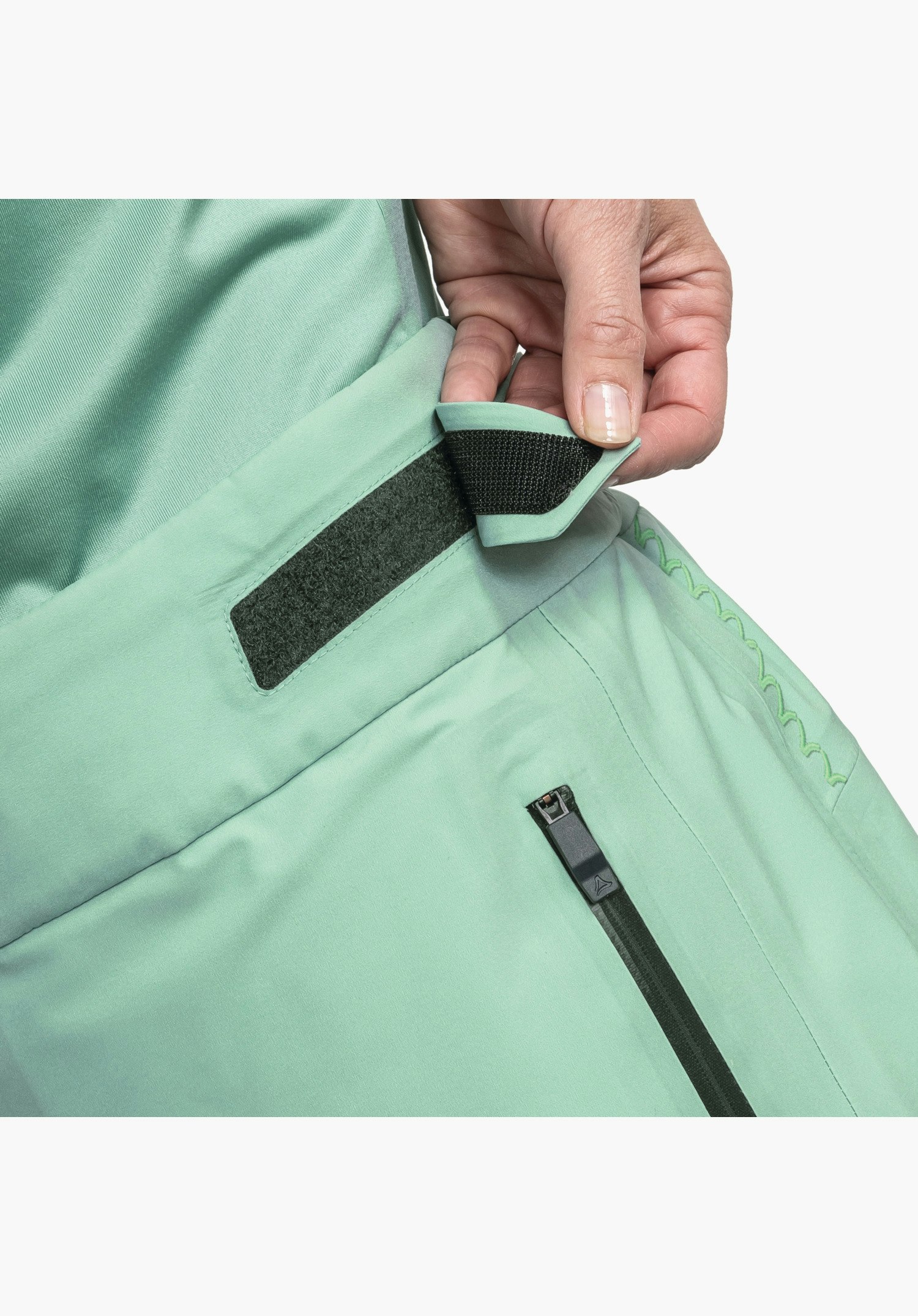 Pants Style Lufeld WMS zeigt den verstellbaren Bund mit Klettverschluss in Grün sowie die Reißverschlusstasche mit Logo-Detail am Zipper, Nahaufnahme vor weißem/neutralem Hintergrund.