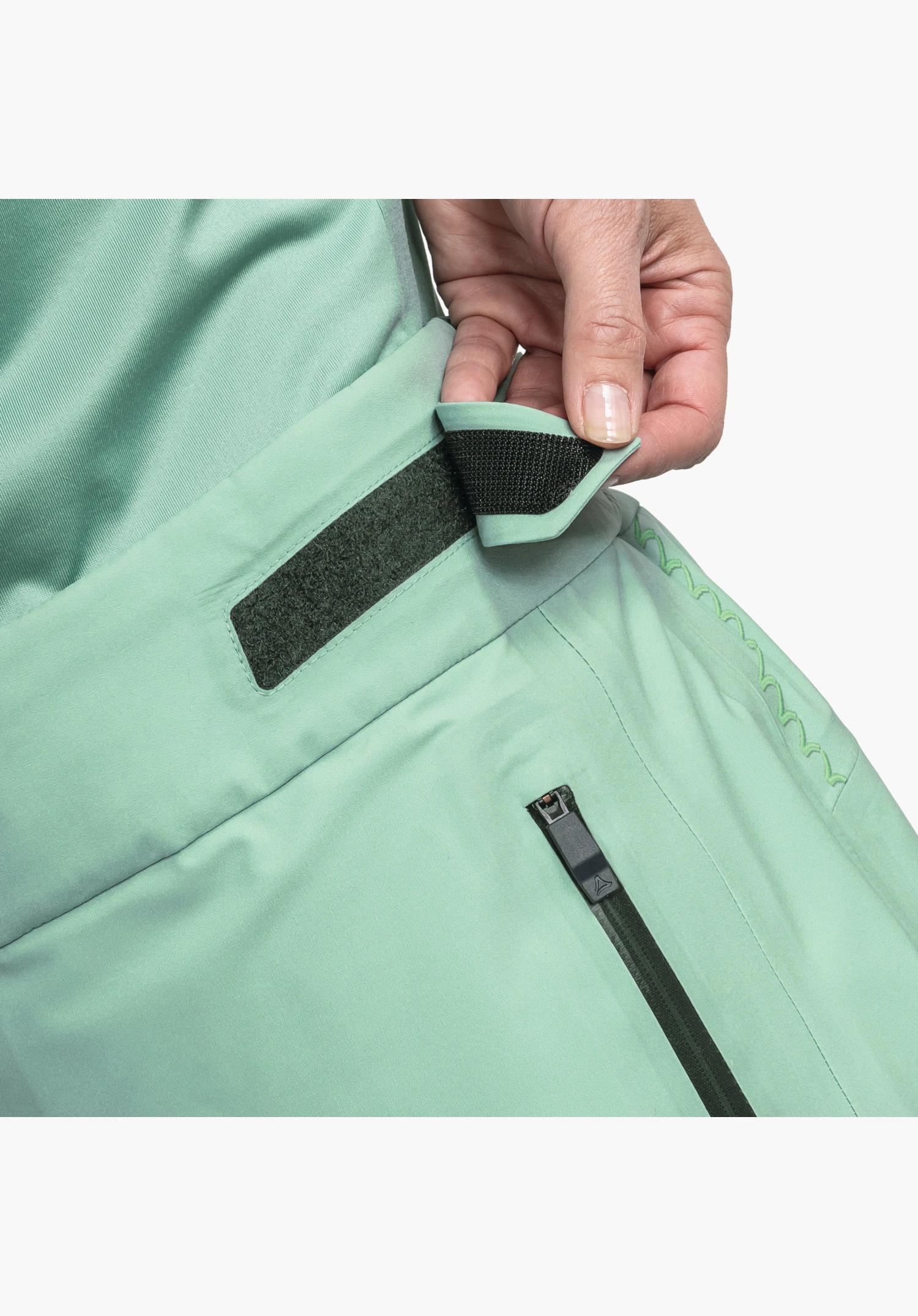 Pants Style Lufeld WMS zeigt den verstellbaren Bund mit Klettverschluss in Grün sowie die Reißverschlusstasche mit Logo-Detail am Zipper, Nahaufnahme vor weißem/neutralem Hintergrund.