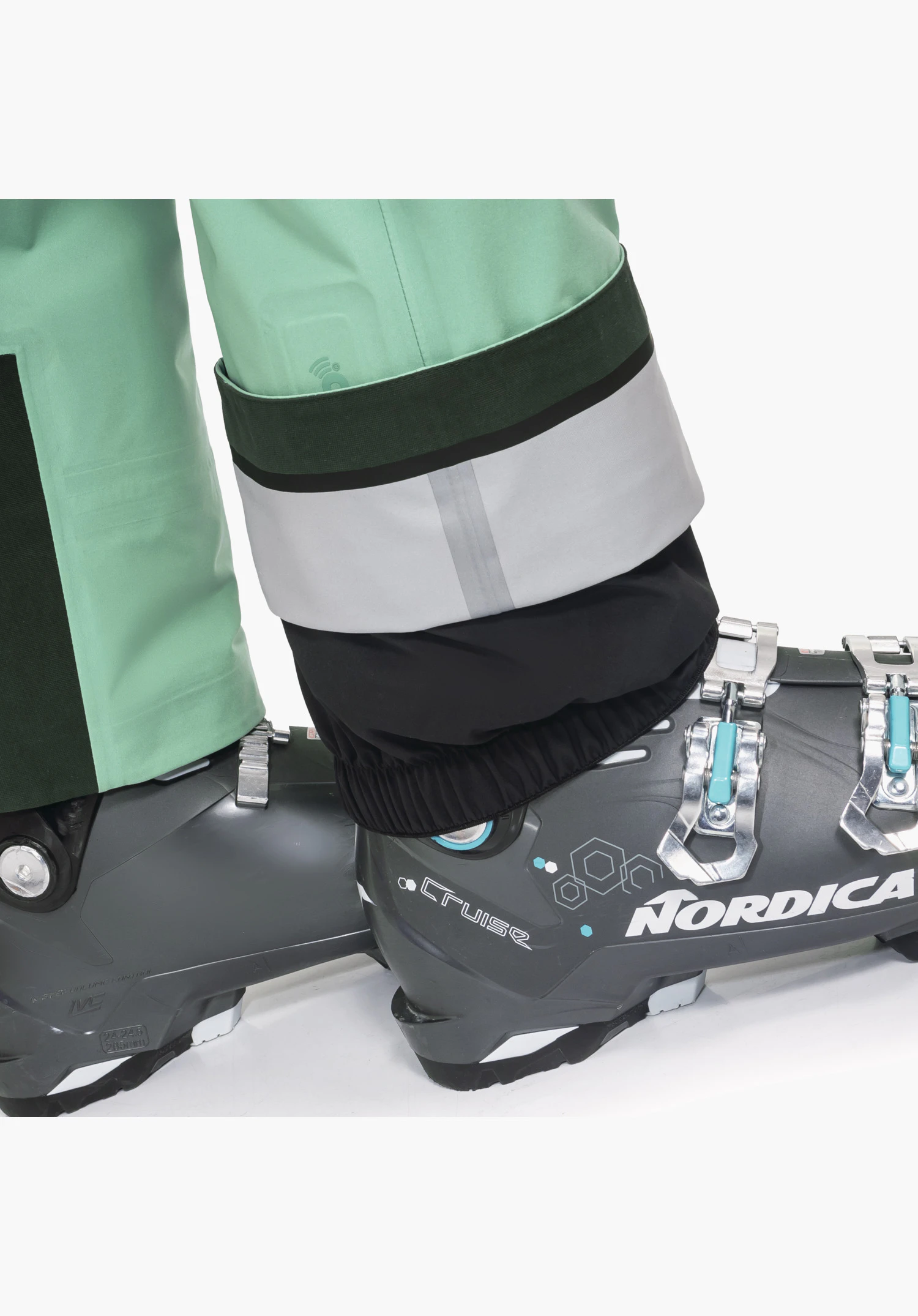 Pants Style Lufeld WMS Detailansicht des grünen Beinabschlusses mit verstärktem Kantenschutz, RECCOÂ®-Reflektor und fixiertem Schneefang am Skischuh, Nahaufnahme vor weißem/neutralem Hintergrund.
