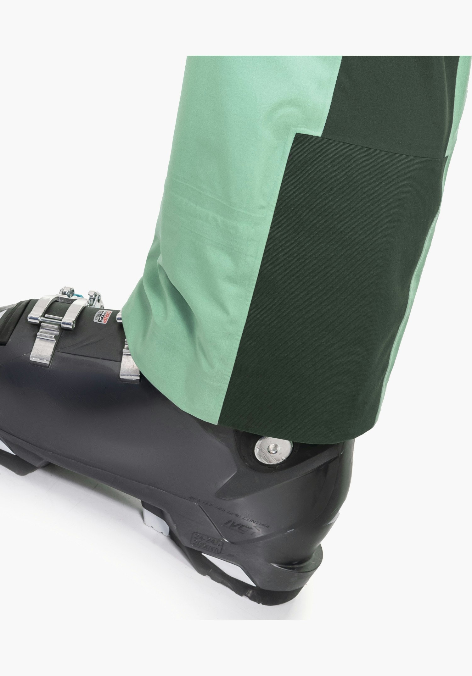 Pants Style Lufeld WMS zeigt den grünen Beinabschluss mit einem robusten, dunkler abgesetzten Kantenschutz über einem Skischuh, Nahaufnahme vor weißem Hintergrund.