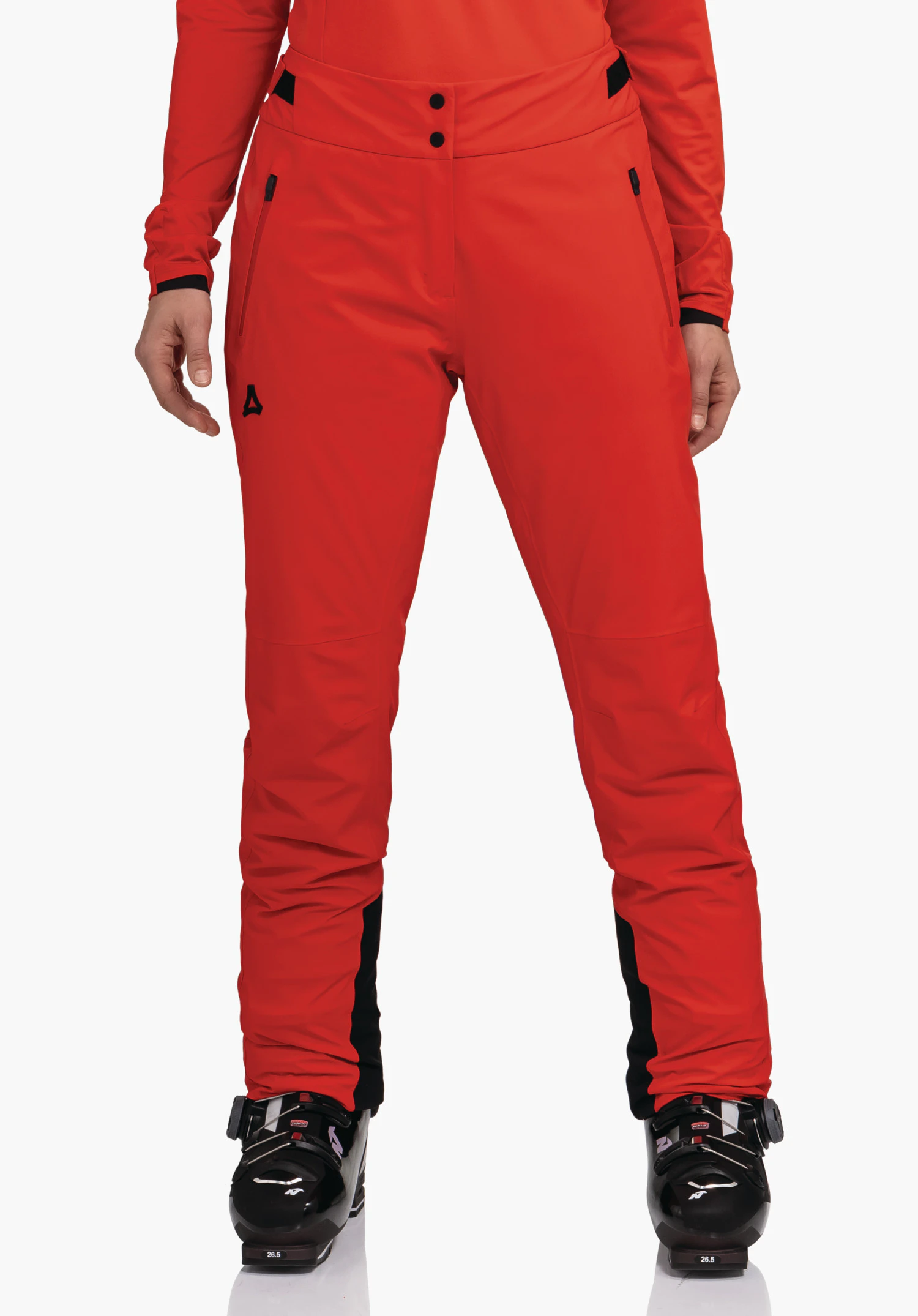 Pants Style Skayra WMS pantalon de ski rouge avec logo Schöffel sur la cuisse, porté par une personne debout; environnement studio blanc, chaussures de ski noires visibles.