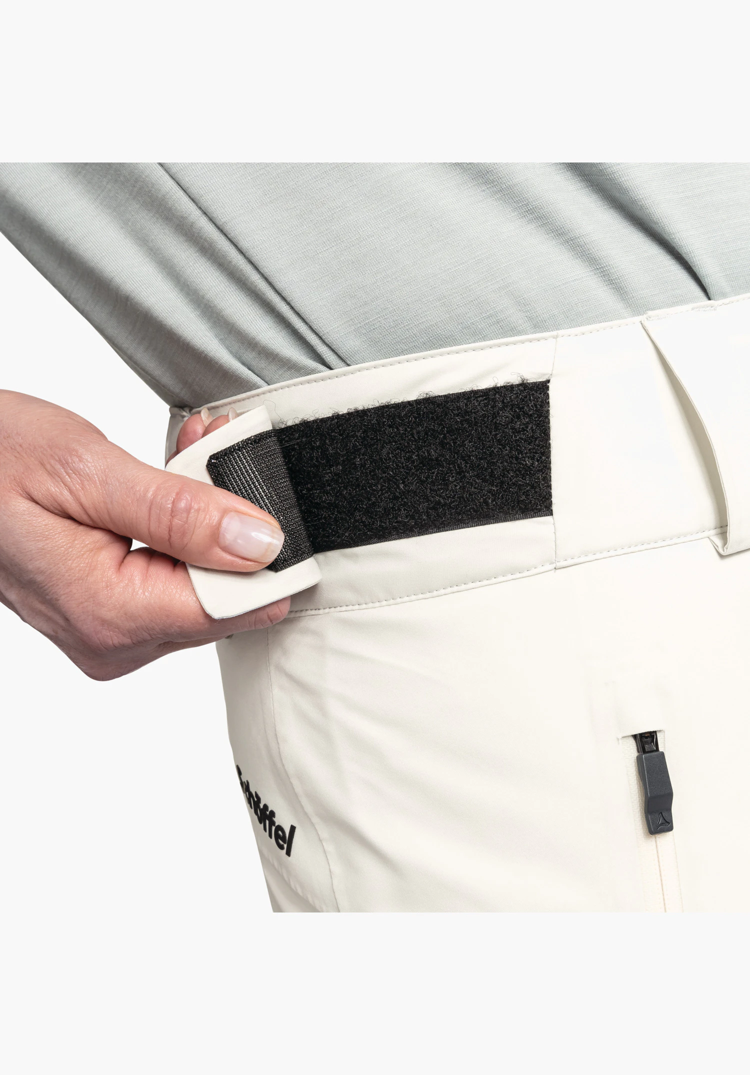 Pants Style Pine WMS detail van de verstelbare tailleband in de kleur natuur. Een zwarte klittenbandsluiting voor wijdteverstelling wordt getoond, met daaronder een rits en het gedeeltelijk zichtbare Schöffel-opschrift. Close-up voor een witte/neutrale achtergrond.