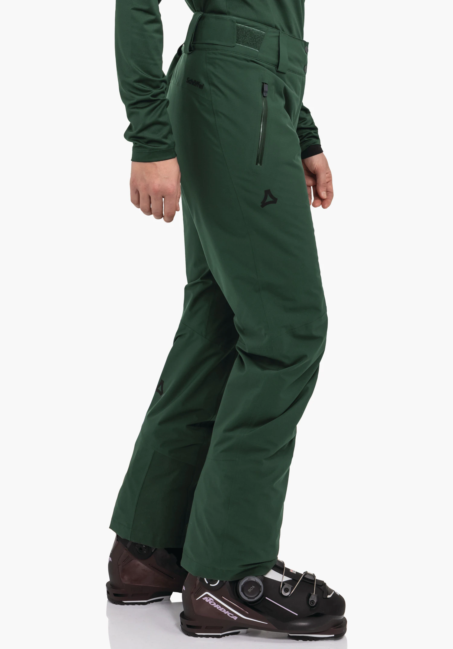 Pants Style Pine WMS donkergroene skibroek, zijwaarts gedragen; persoon staat stil; broek met ritszak en Schöffel-logo, tekst "Schöffel" op de heup; zwarte Nordica-skischoenen zichtbaar; witte achtergrond.