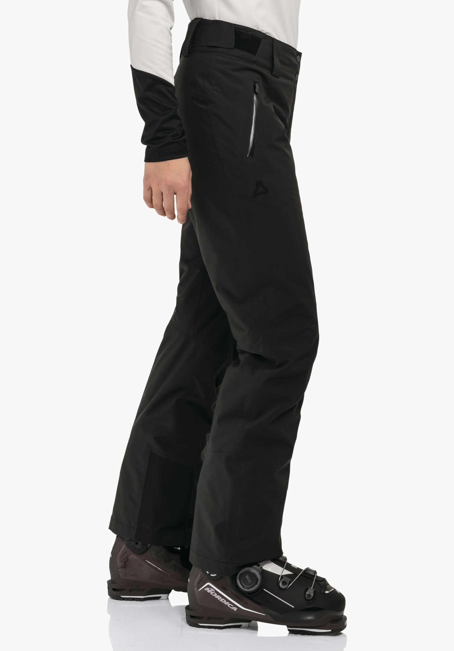 Pants Style Pine WMS pantalon de ski noir avec logo Schöffel; porté par une personne debout devant un fond blanc, combiné avec un haut blanc et des chaussures de ski noires avec inscriptions «NORDICA», «BOA».