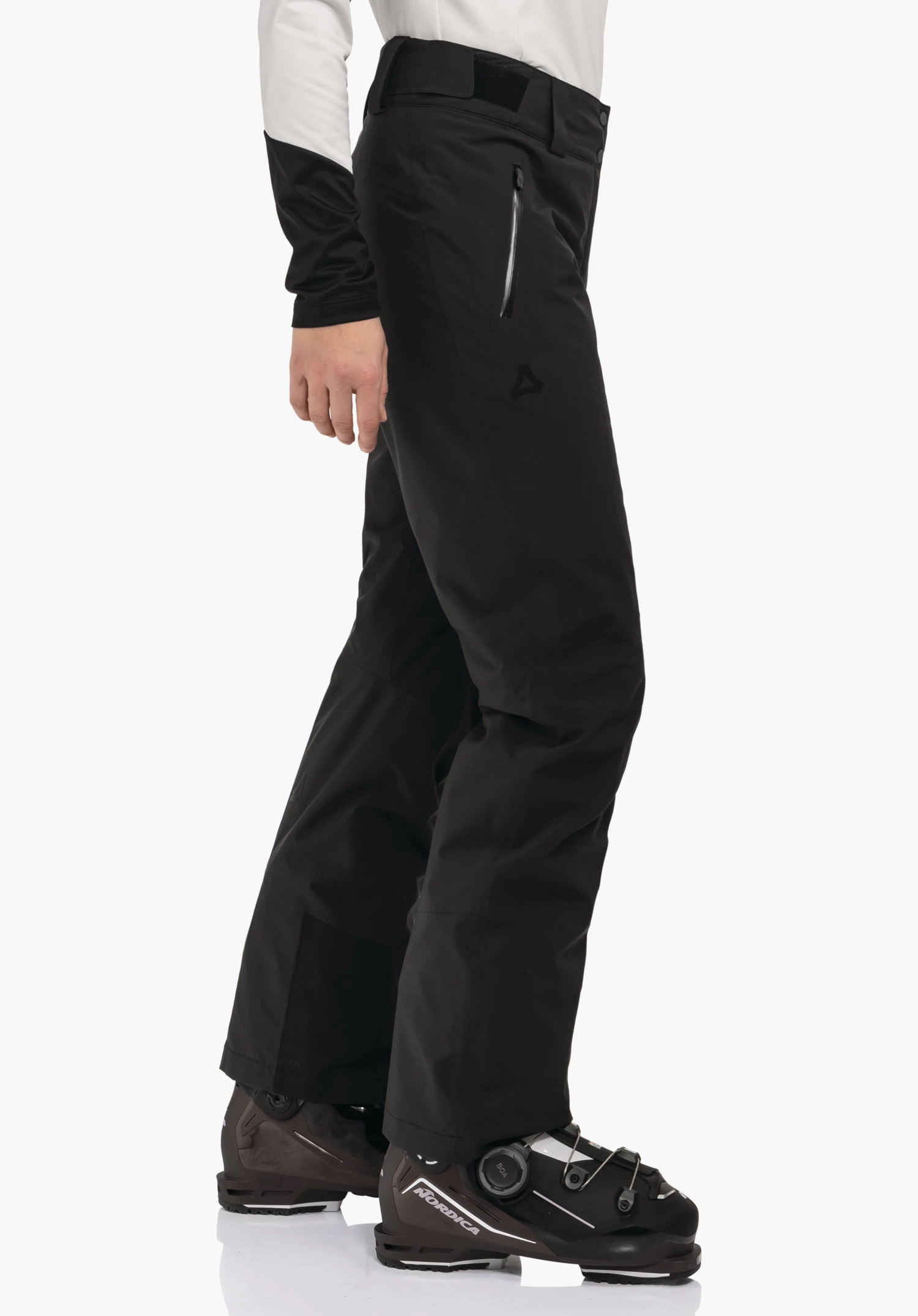 Pants Style Pine WMS zwarte skibroek met Schöffel-logo, gedragen door een staande persoon op een witte achtergrond, gecombineerd met een wit bovenstuk en zwarte skischoenen met de teksten NORDICA, BOA.