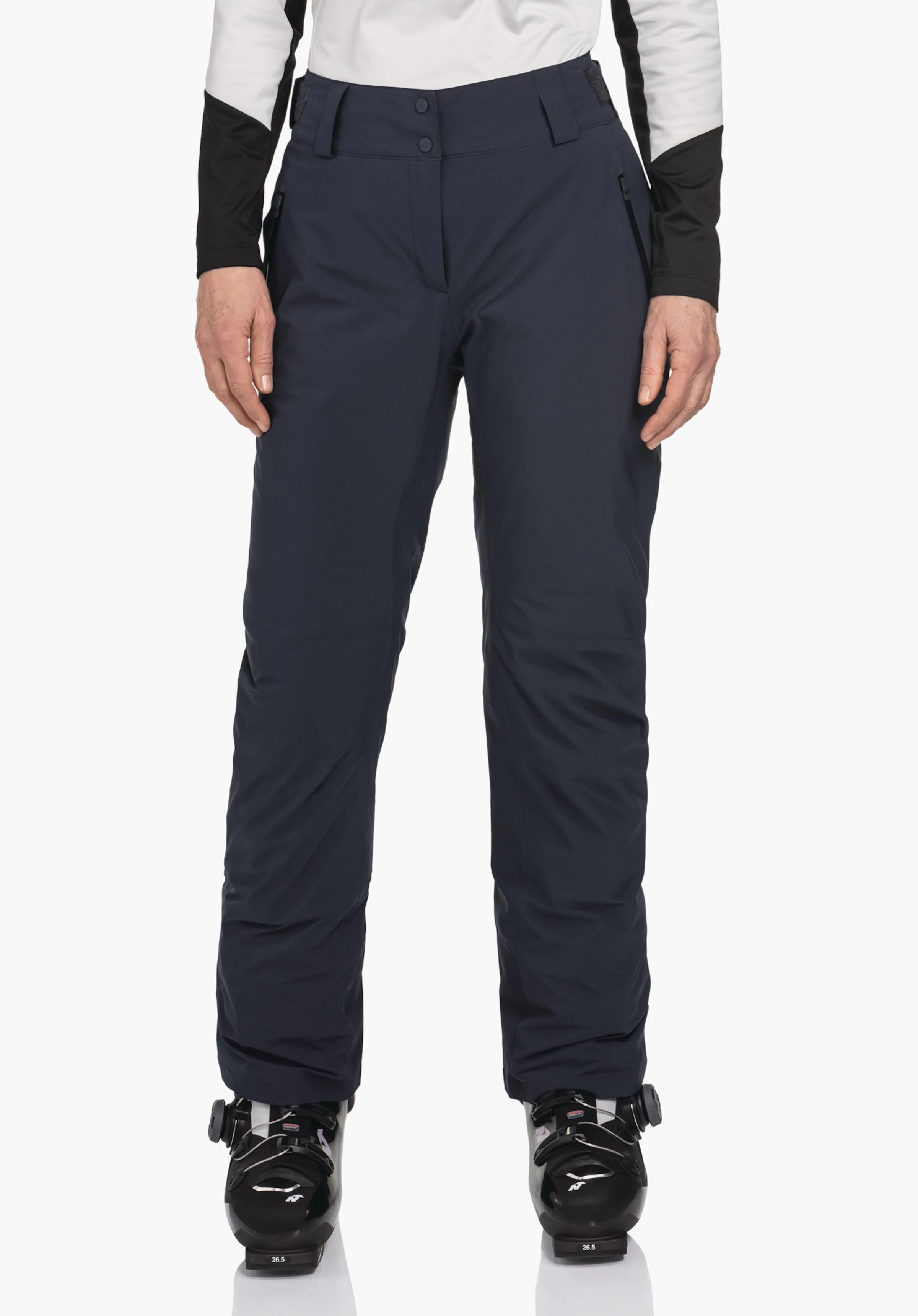 Pants Style Pine WMS pantalon de ski bleu foncé, porté par une personne debout; bras détendus le long du corps, haut blanc avec empiècements noirs, chaussures de ski noires; fond blanc neutre.