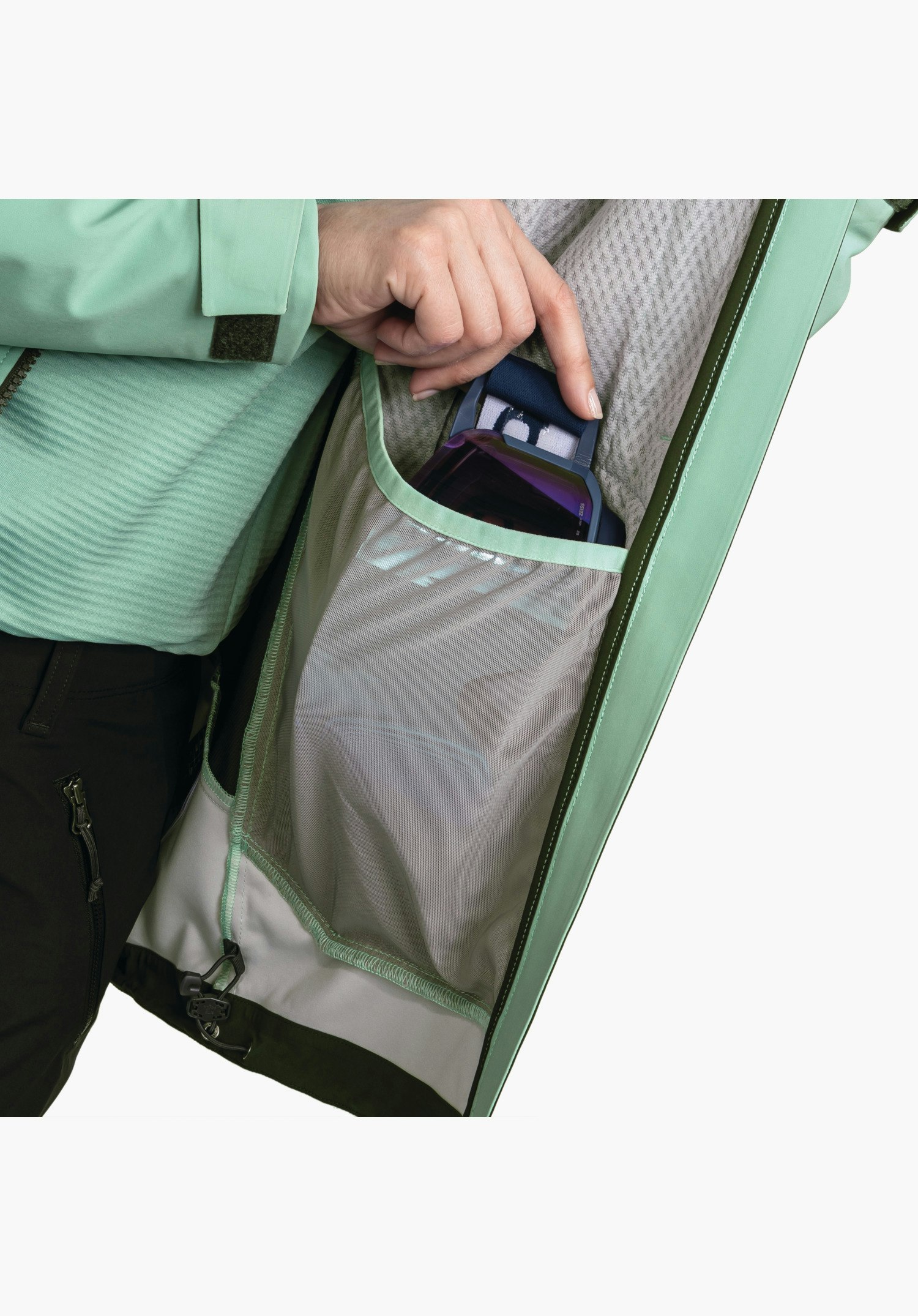 Softshell Jk Style Milagle WMS mit Blick auf das Innenfutter der grünen Jacke, das eine praktische Netztasche für die Skibrille zeigt, in die gerade eine Hand greift. Detailaufnahme vor weißem/neutralem Hintergrund.