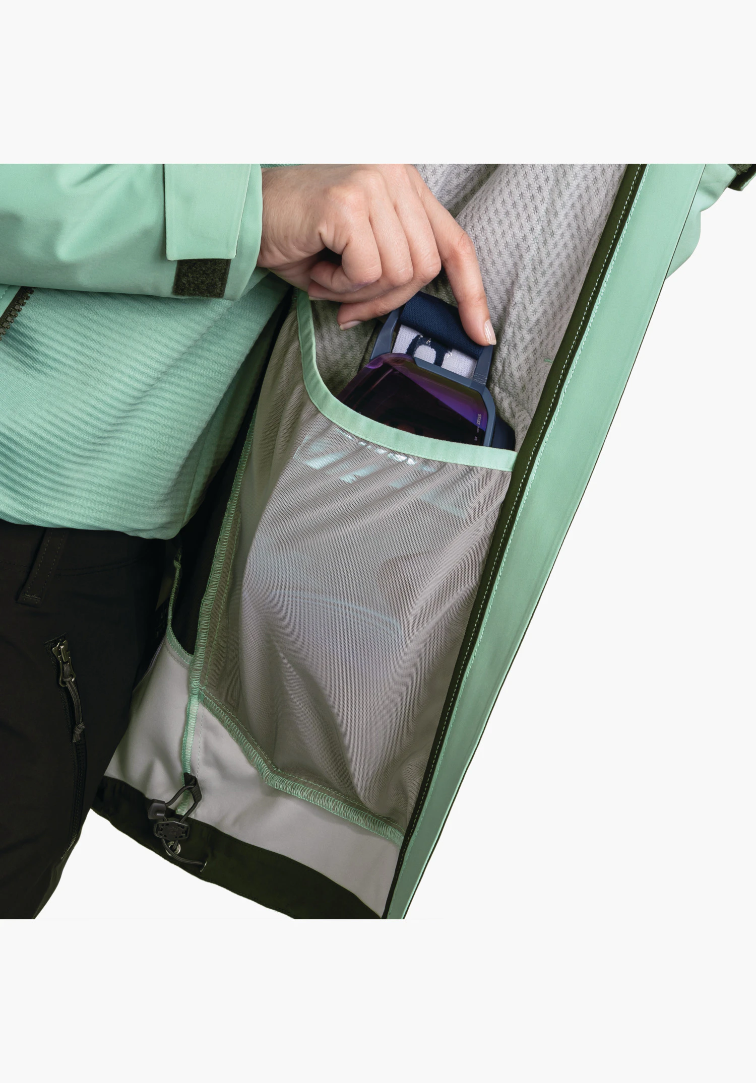 Softshell Jk Style Milagle WMS montrant la doublure de la veste verte, dotée d'une poche en filet pratique pour les lunettes de ski, dans laquelle une main s'introduit. Gros plan sur fond blanc/neutre.