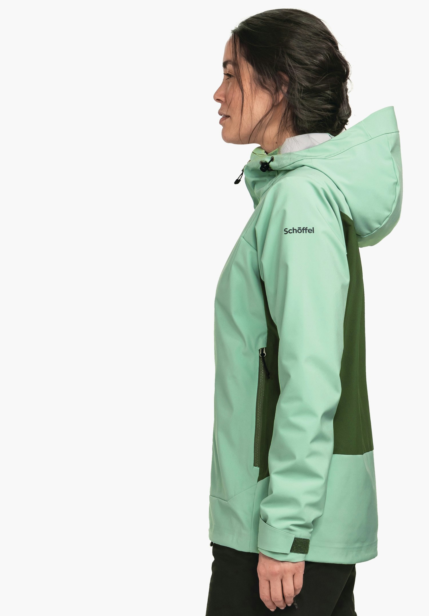 Softshell Jk Style Milagle WMS Frau trägt hellgrüne Jacke mit Kapuze und dunkelgrünen Seiteneinsätzen, steht seitlich vor weißem Hintergrund; auf Ärmel steht "Schöffel".
