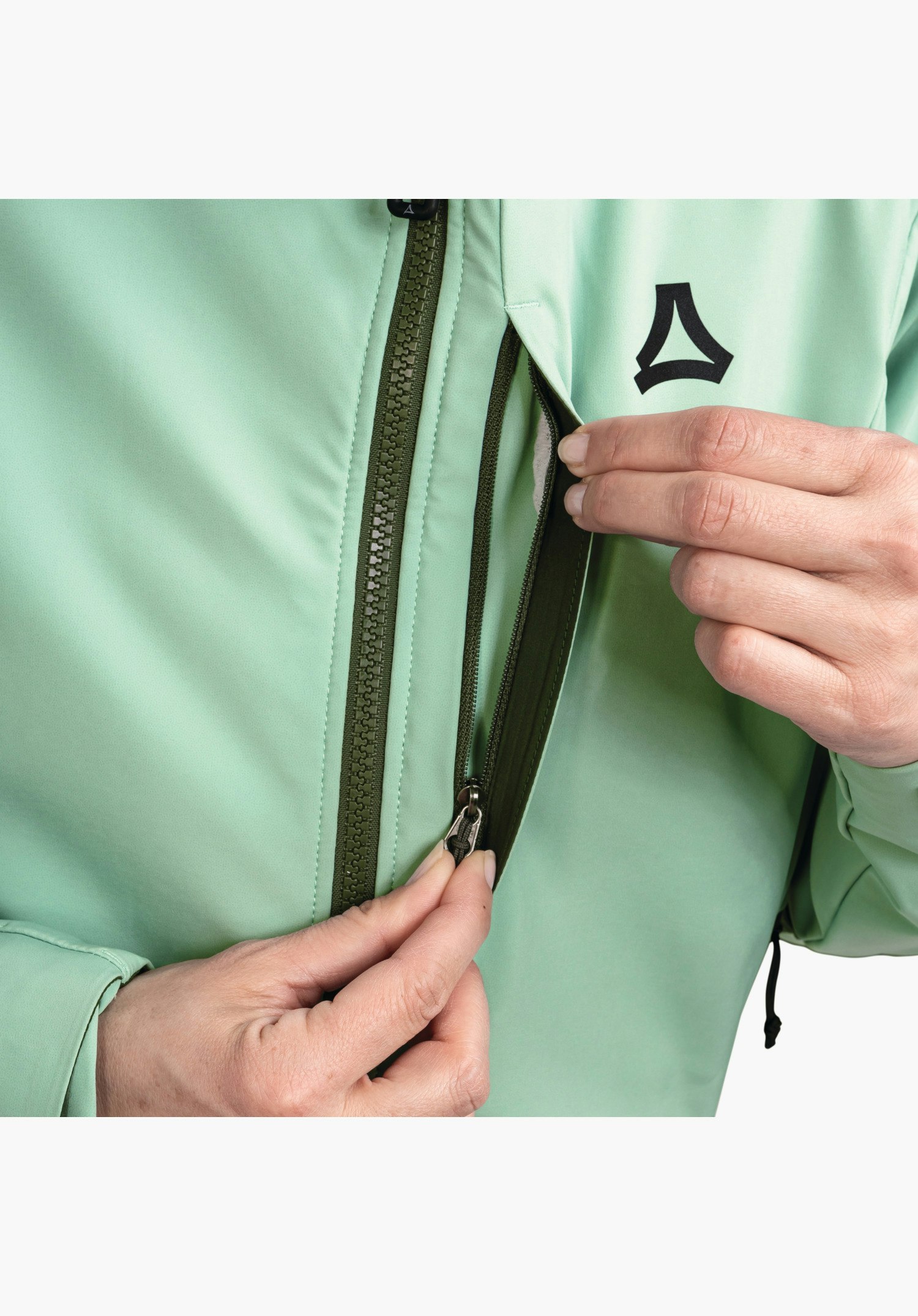 Softshell Jk Style Milagle WMS zeigt die Brusttasche mit Reißverschluss, den dunkelgrünen Frontreißverschluss und das Schöffel-Logo auf grünem Stoff. Nahaufnahme vor weißem Hintergrund.