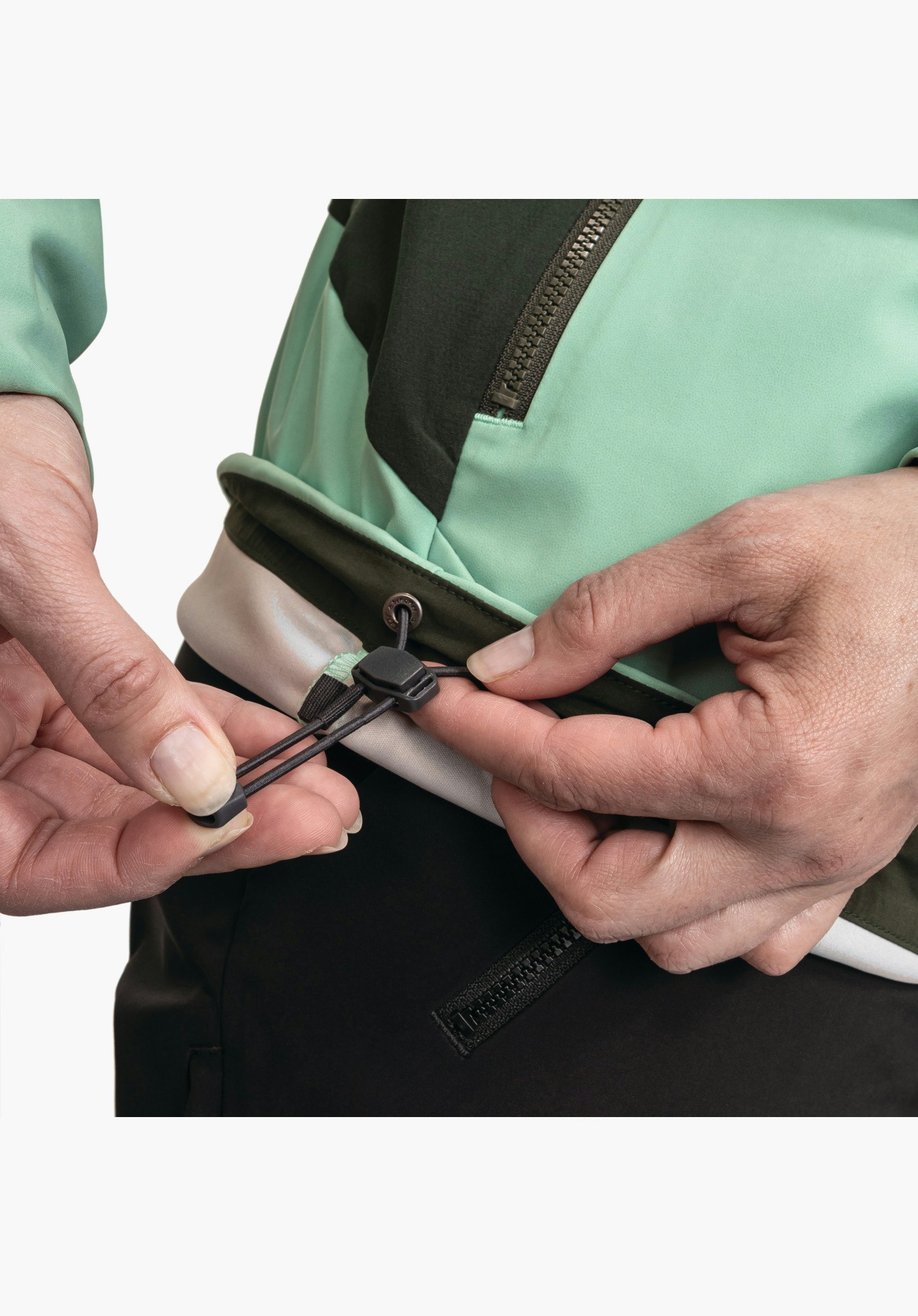 Softshell Jk Style Milagle WMS, Detailansicht der Hände beim Anpassen des verstellbaren Saums mit elastischem Kordelzug und Stopper an der grünen Jacke, Nahaufnahme vor weißem/neutralem Hintergrund.