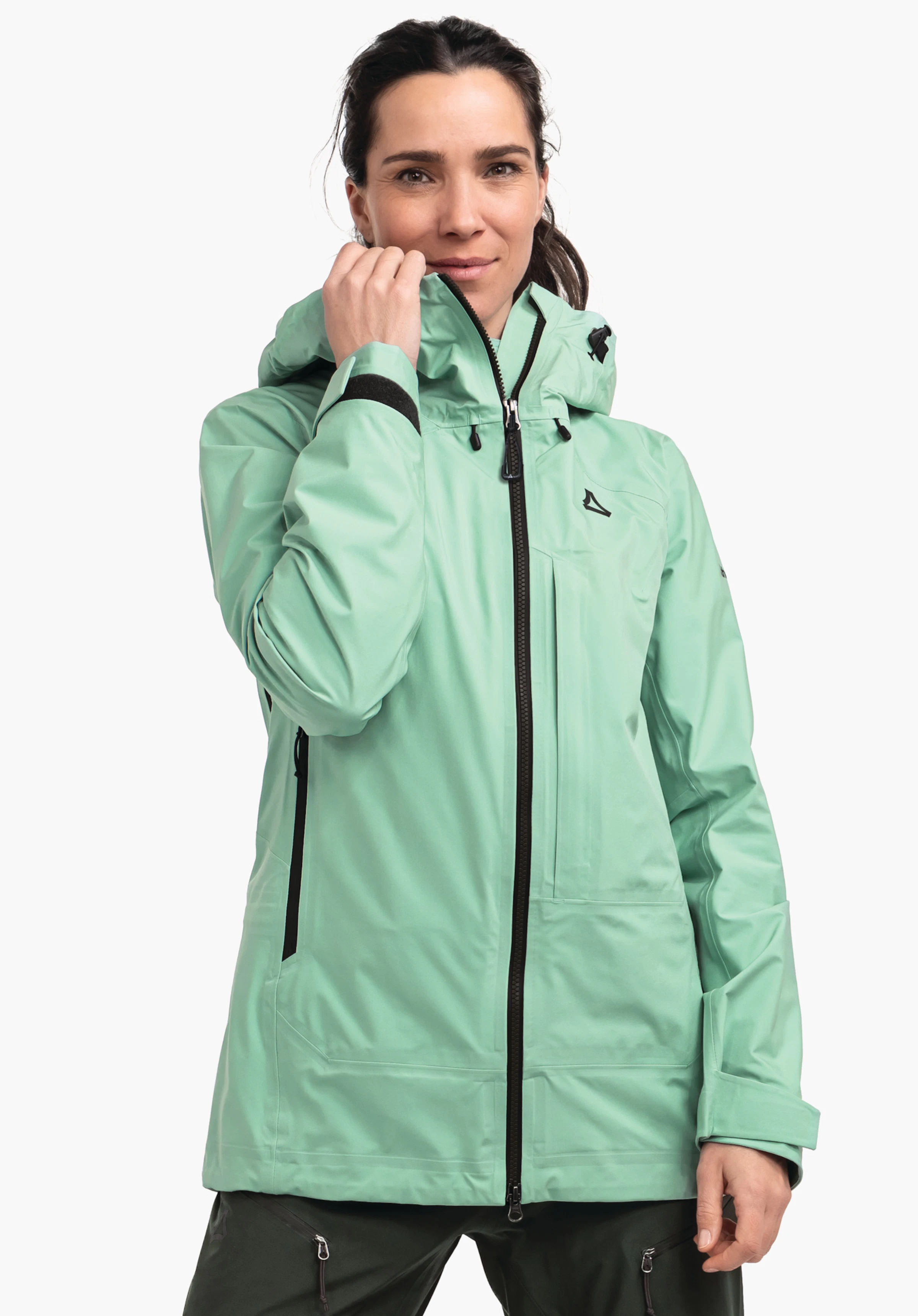 Jacket Style Milagle WMS lichtgroene outdoorjas; vrouw trekt de rits bij de kraag omhoog, Schöffel-logo op de borst, zwarte ritsen en details, capuchon; staande opname voor een witte achtergrond.