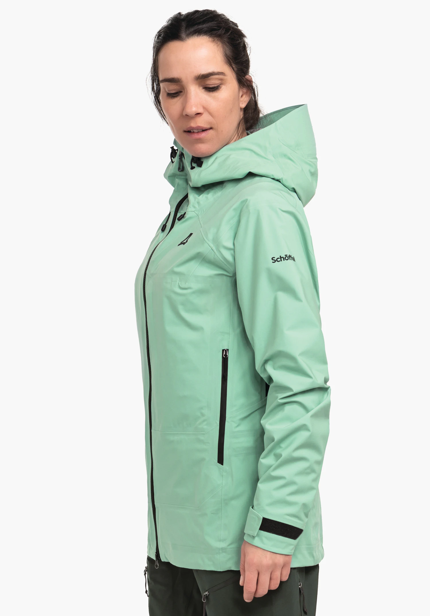 Jacket Style Milagle WMS veste: personne porte une veste de pluie vert clair longueur hanches avec capuche et fermetures éclair noires; regarde vers le bas de profil devant fond blanc. Texte: "Schöffel". Logo Schöffel (triangulaire) visible.