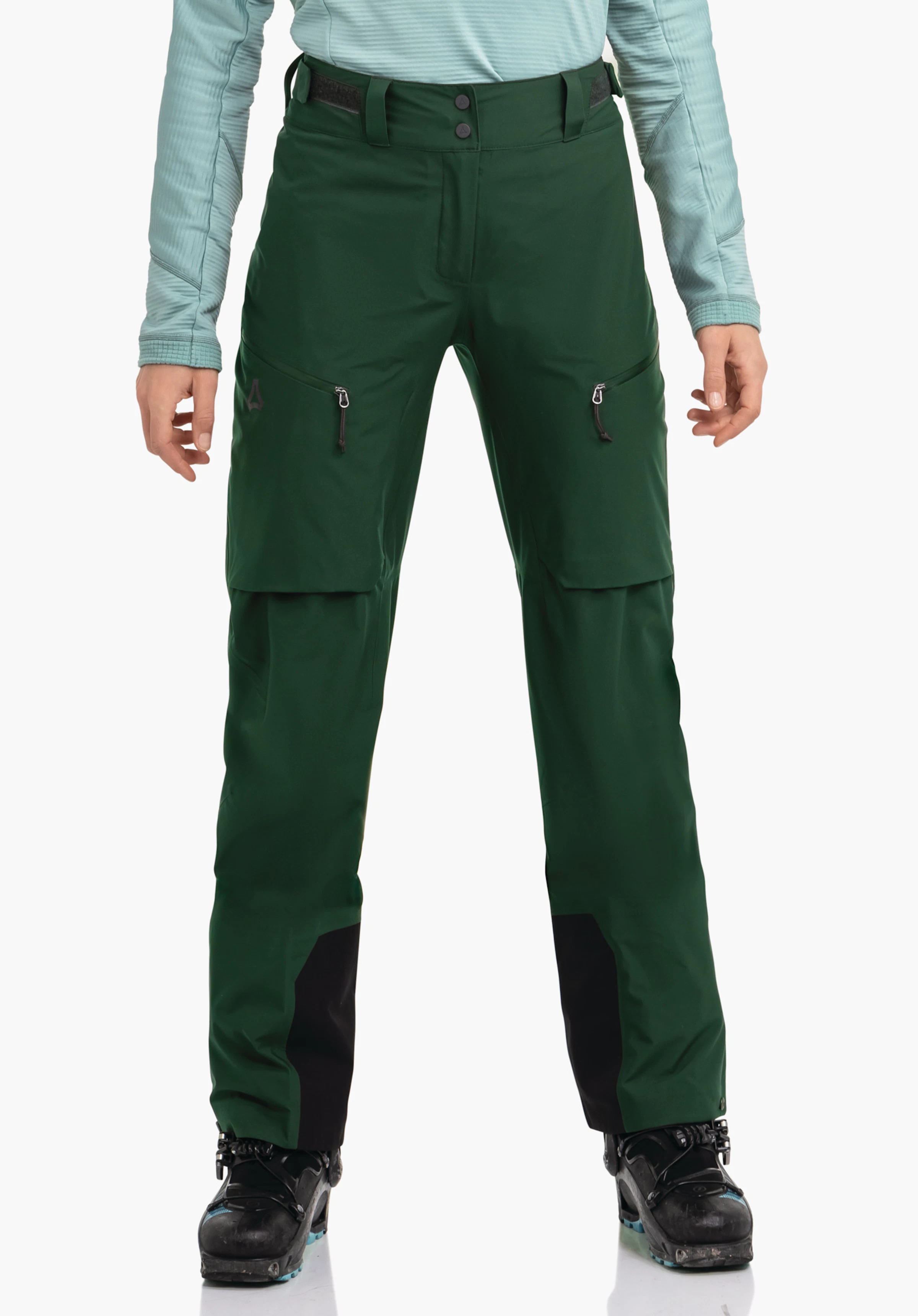 Pants Style Milagle WMS dunkelgrüne Skihose, sitzt an der Hüfte einer stehenden Person; zwei Reißverschlusstaschen, schwarze Beinverstärkungen, Schöffel-Logo am linken Oberschenkel; hellblaues Oberteil, weißer Hintergrund.