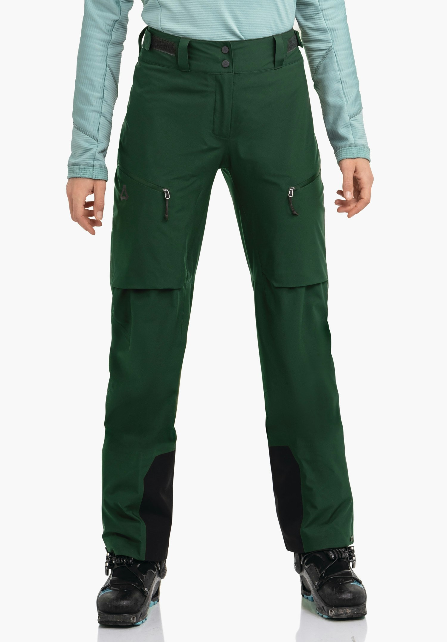 Pants Style Milagle WMS pantalon de ski vert foncé porté à la taille par une personne debout; deux poches zippées, renforts noirs aux jambes, logo Schöffel sur la cuisse gauche; haut bleu clair, fond blanc.
