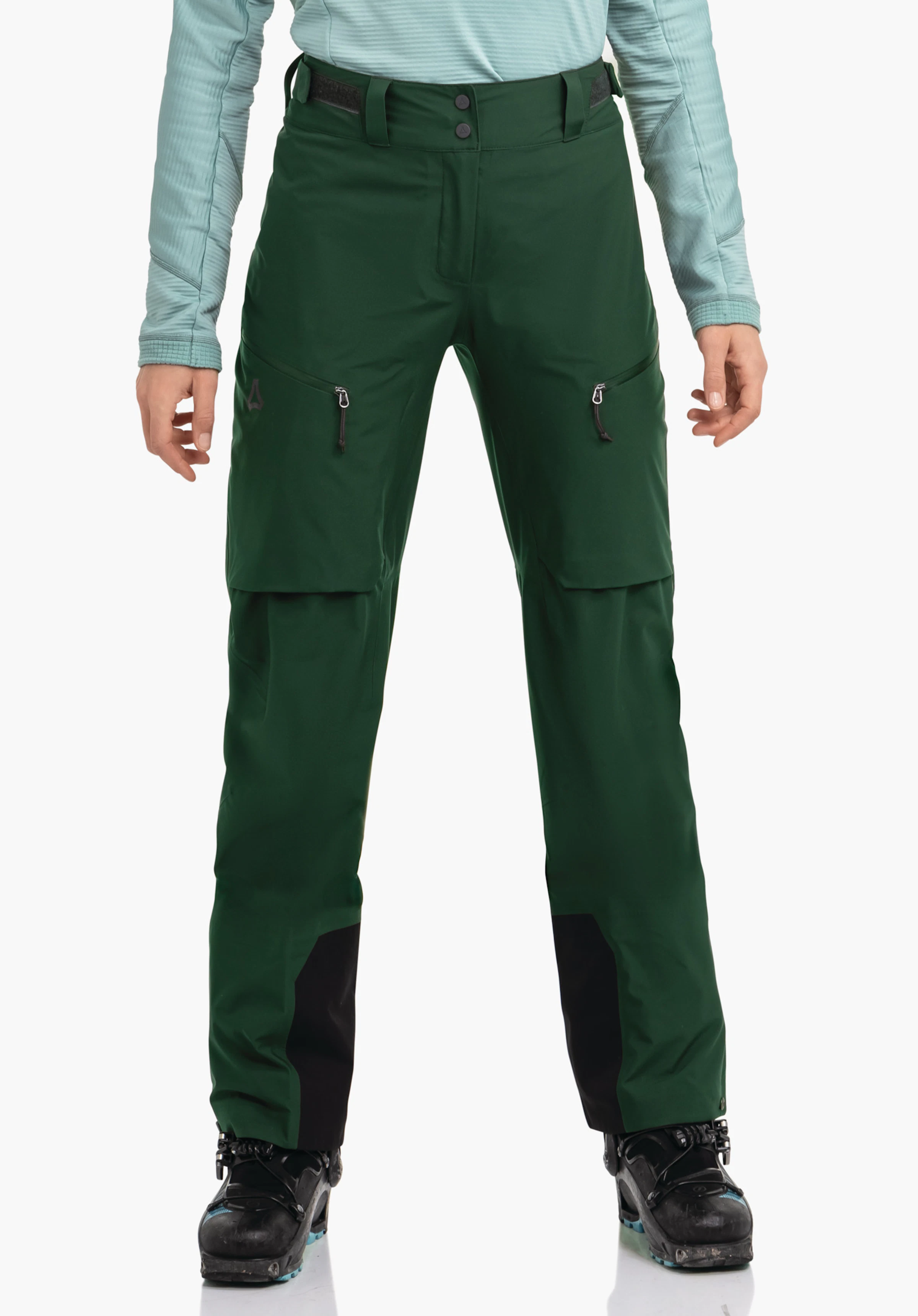 Pants Style Milagle WMS dunkelgrüne Skihose, sitzt an der Hüfte einer stehenden Person; zwei Reißverschlusstaschen, schwarze Beinverstärkungen, Schöffel-Logo am linken Oberschenkel; hellblaues Oberteil, weißer Hintergrund.