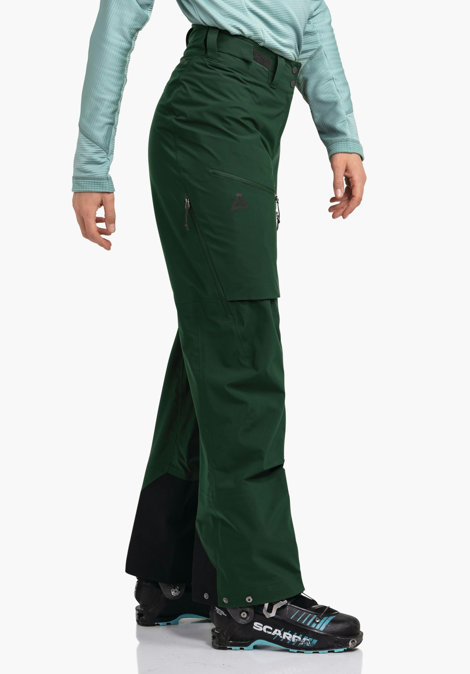 Pants Style Milagle WMS Dunkelgrüne Outdoorhose mit Schöffel-Logo, seitlich getragen, Bein leicht angewinkelt; Kontext: hellblaues Langarmshirt, weiße Studiofläche, schwarze Skistiefel mit hellblauer Sohle. Text: "SCARPA"