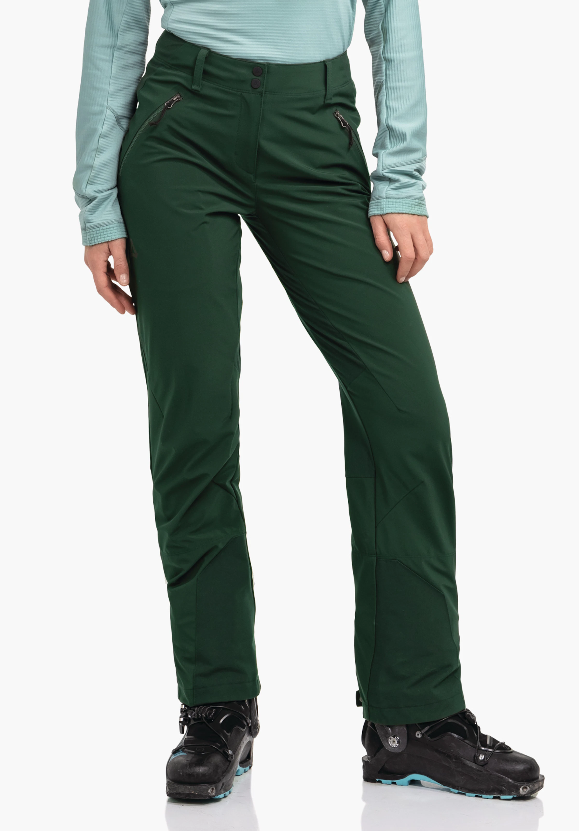 Softshell Pt Style Milagle WMS Dunkelgrüne Skihose, stehend getragen, zeigt Reißverschlusstaschen und Schöffel-Logo; Person steht vor weißem Hintergrund, trägt hellblaues Oberteil und schwarze Skischuhe.