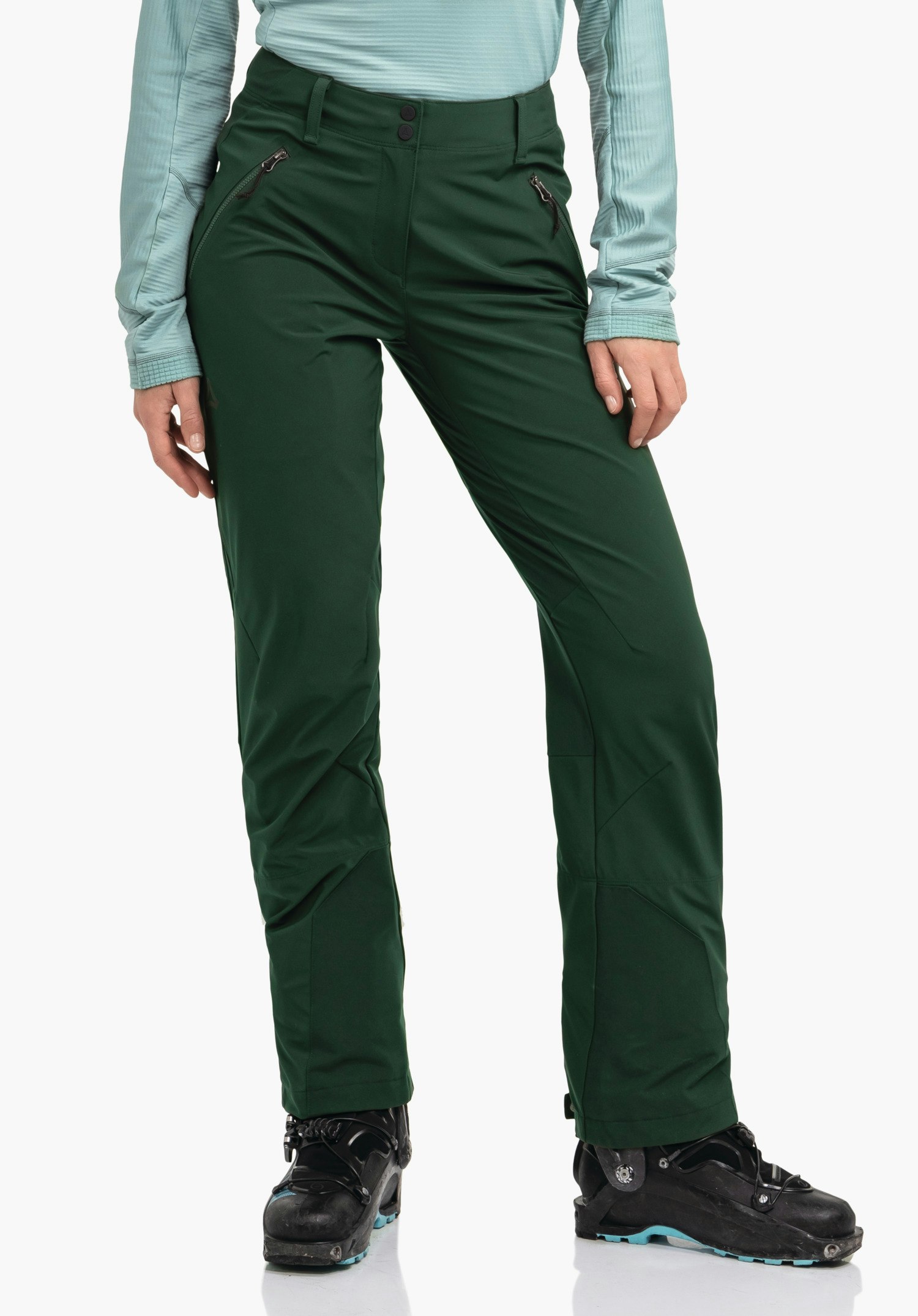 Softshell Pt Style Milagle WMS pantalon de ski vert foncé porté debout, montrant des poches zippées et le logo Schöffel; personne debout devant un fond blanc, portant un haut bleu clair et des chaussures de ski noires.