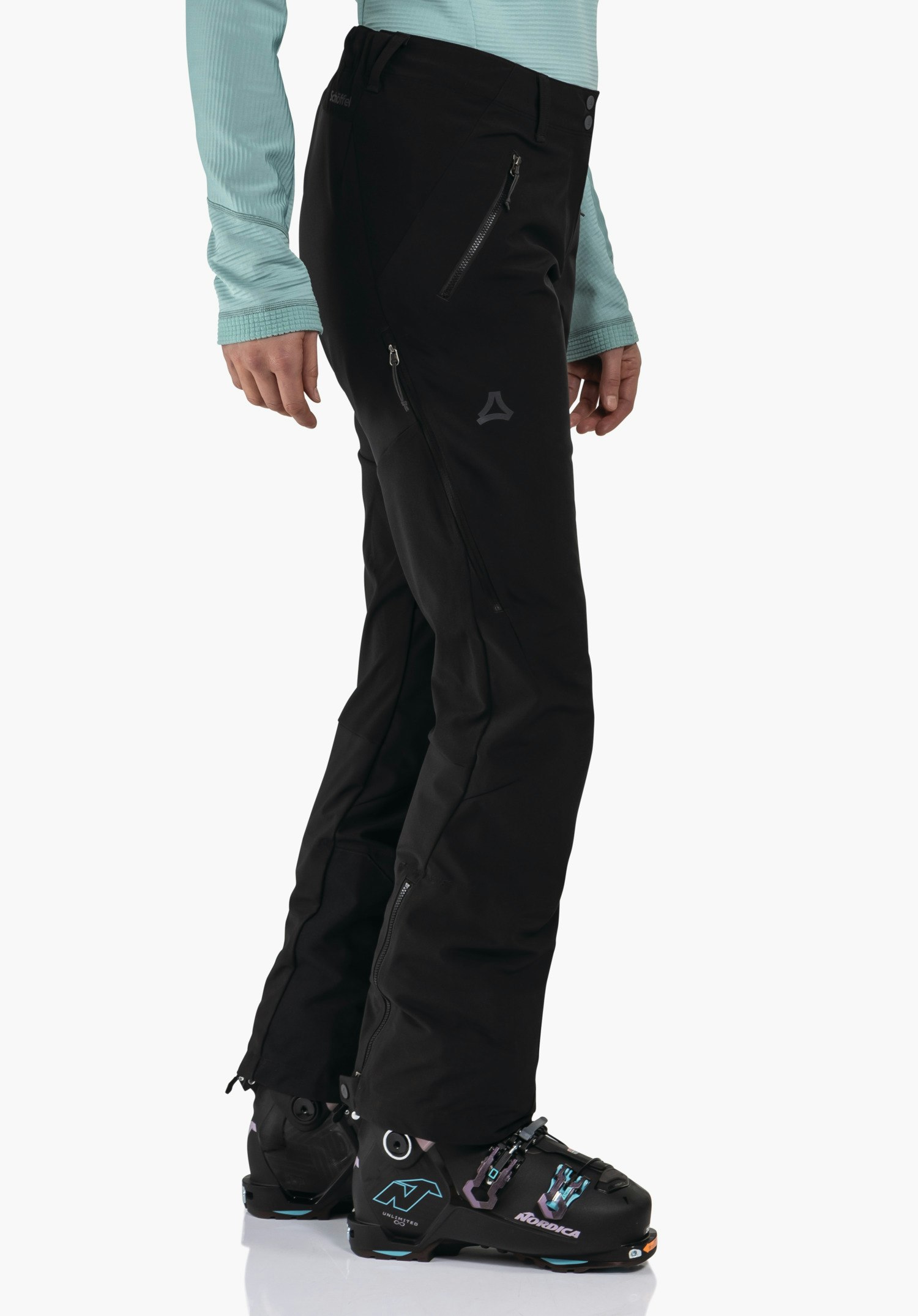 Softshell Pt Style Milagle WMS pantalon de ski noir avec logo Schöffel, porté par un modèle debout; combiné avec un haut turquoise clair et des chaussures de ski noires, fond blanc. Texte: Schöffel, NORDICA.