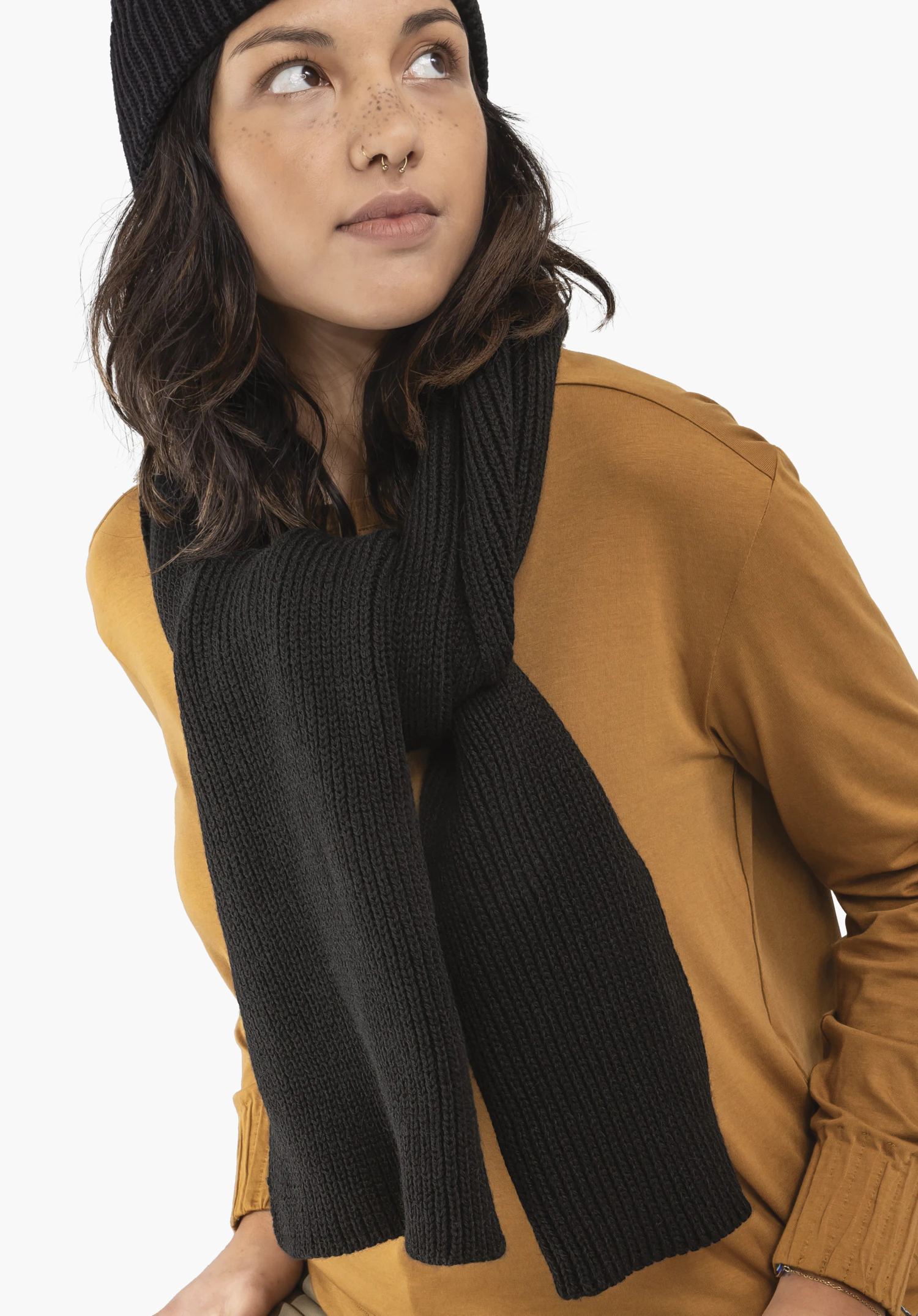 Scarf Style Boslix ALL écharpe tricotée noire drapée lâchement autour du cou ; personne portant un pull orange et un bonnet noir, regarde vers le haut, fond blanc.
