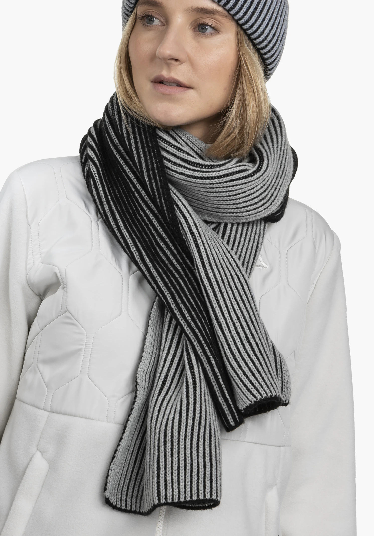 Scarf Style Cledio ALL gestreepte sjaal in zwart en lichtgrijs om de hals gewikkeld; vrouw draagt een bijpassende muts, witte gewatteerde jas met Schöffel-logo, neutrale lichte achtergrond.