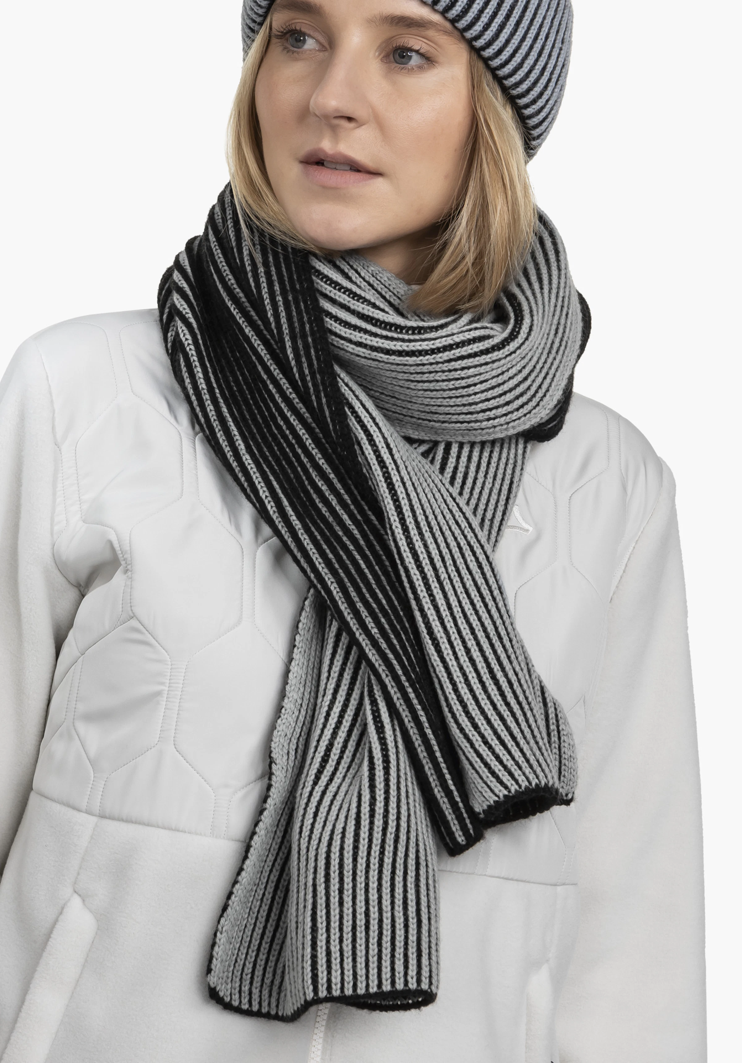 Scarf Style Cledio ALL gestreepte sjaal in zwart en lichtgrijs om de hals gewikkeld; vrouw draagt een bijpassende muts, witte gewatteerde jas met Schöffel-logo, neutrale lichte achtergrond.