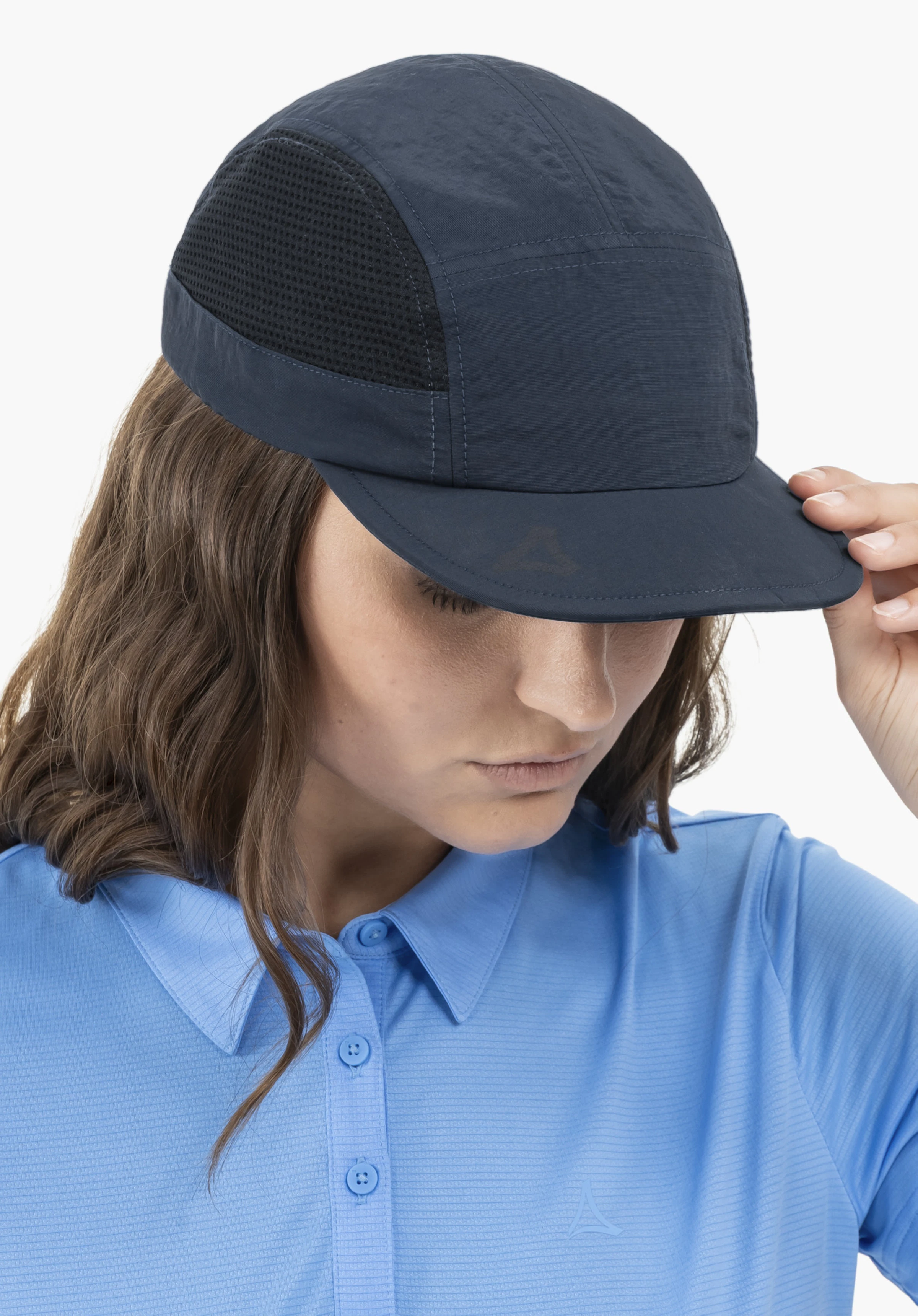 Cap Style Mauku UNI worn by a model looking downwards and holding the brim of the blue cap with her hand, revealing a subtle Schöffel-Logo on the peak. The close-up shot is taken against a neutral background.