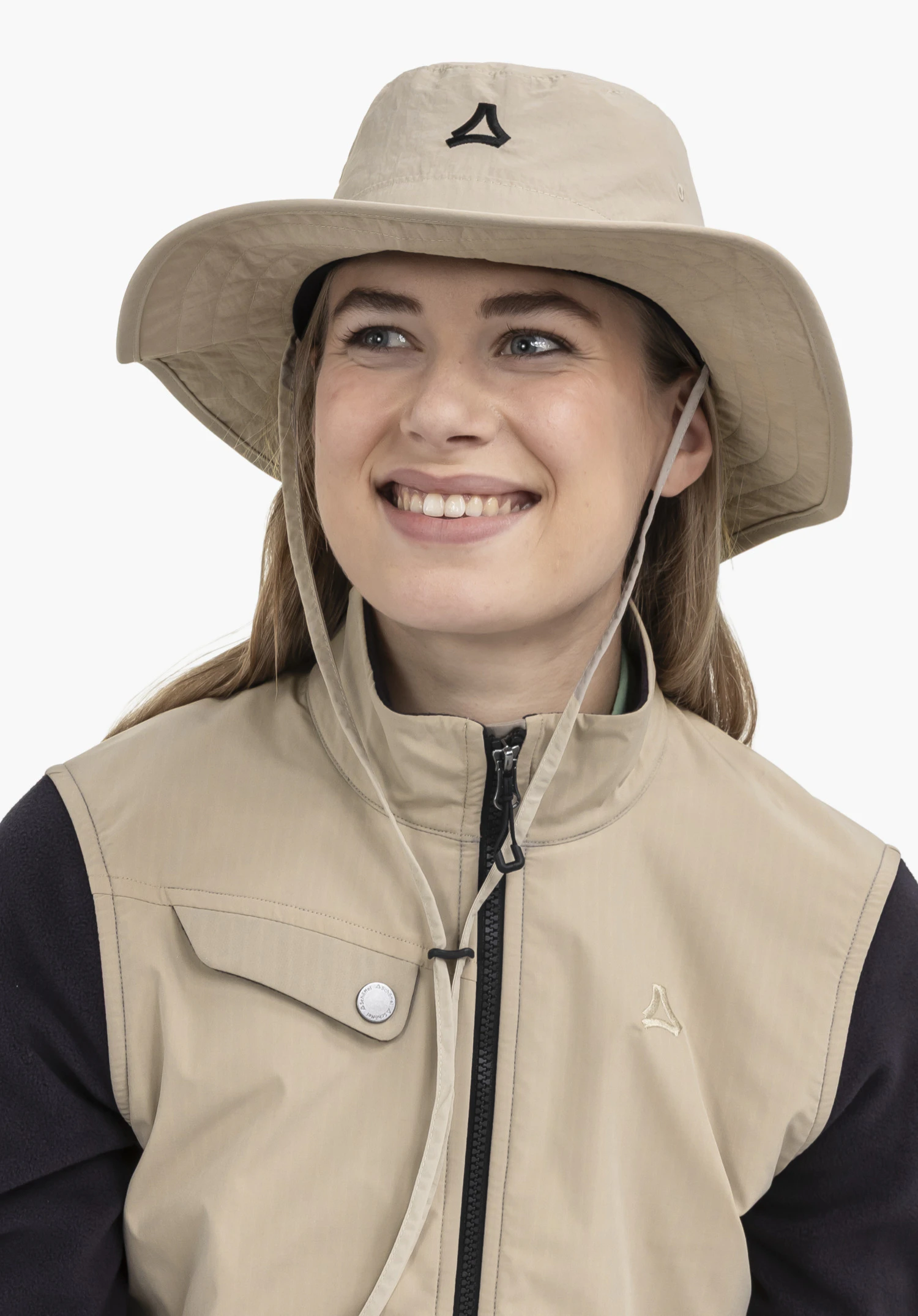 Hat Style Fraydo UNI wird von einem lächelnden Model getragen, das frontal abgebildet ist und den Blick leicht zur Seite wendet. Der beigefarbene Hut ist mit einem praktischen Kinnband ausgestattet und präsentiert ein gut sichtbares, schwarzes Schöffel-Logo auf der Stirnseite. Die Aufnahme zeigt das Accessoire vor einem neutralen Hintergrund.