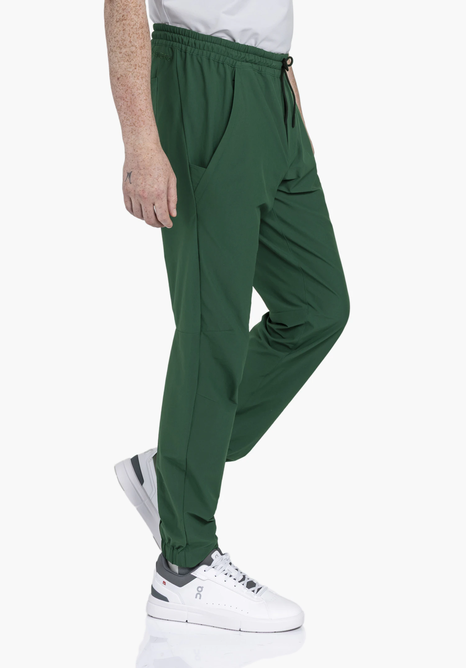 Pants Vienna M en vert, présenté dans une vue latérale sur un fond neutre. Le modèle porte le pantalon à taille élastique avec le logo Schöffel discrètement brodé sur la jambe, dans une pose dynamique.