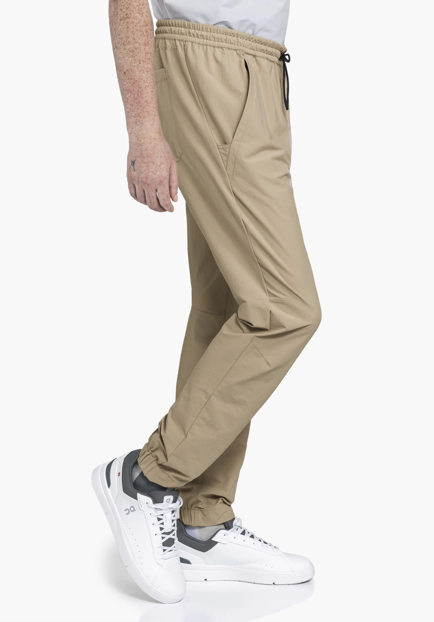 Pants Vienna M in beige: A model is standing sideways against a neutral background, showcasing the trousers with an elasticated waistband. The subtle Schöffel logo on the waistband highlights the clean, everyday aesthetic.
