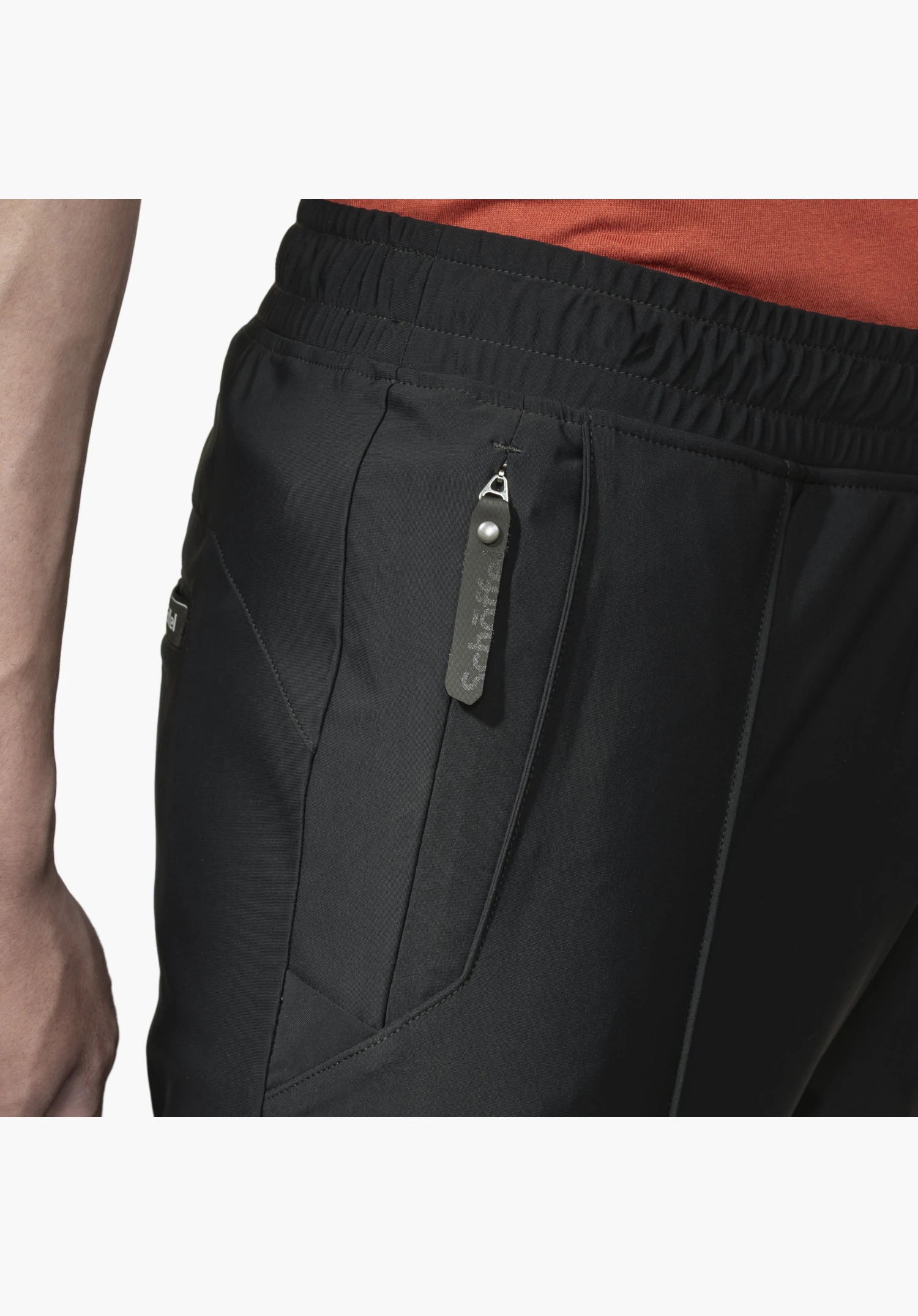 Jersey Pants Style Calmay MEN. Vue détaillée de la poche zippée sur le tissu noir avec une tirette distinctive portant le logo Schöffel, en dessous de la taille élastiquée. Gros plan sur fond blanc/neutre.