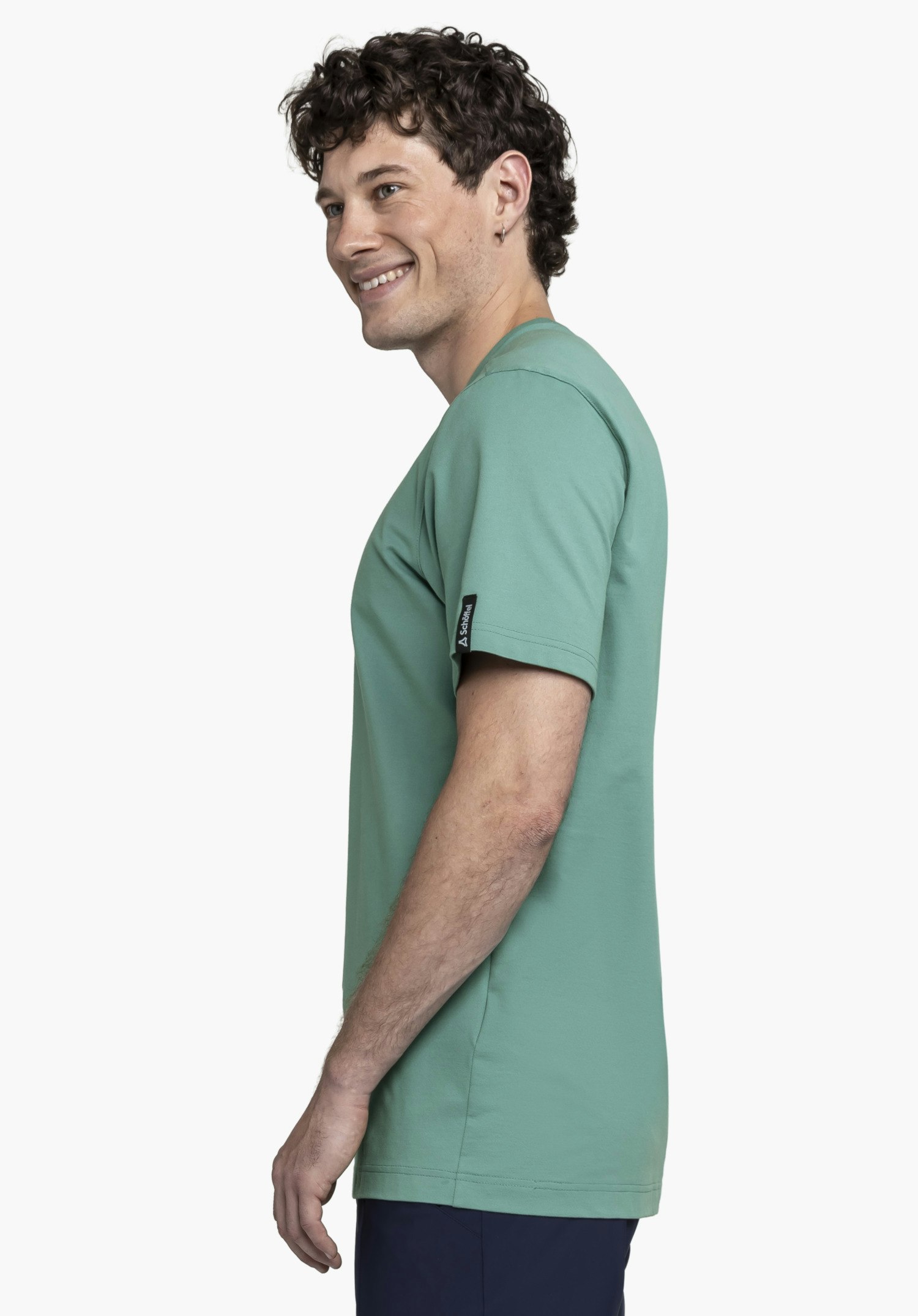 T Shirt Style Serenio MEN präsentiert sich in einem frischen Grün. Das Model steht seitlich vor einem neutralen Hintergrund und lächelt, wobei das Schöffel-Logo unauffällig am Ärmelsaum platziert ist.