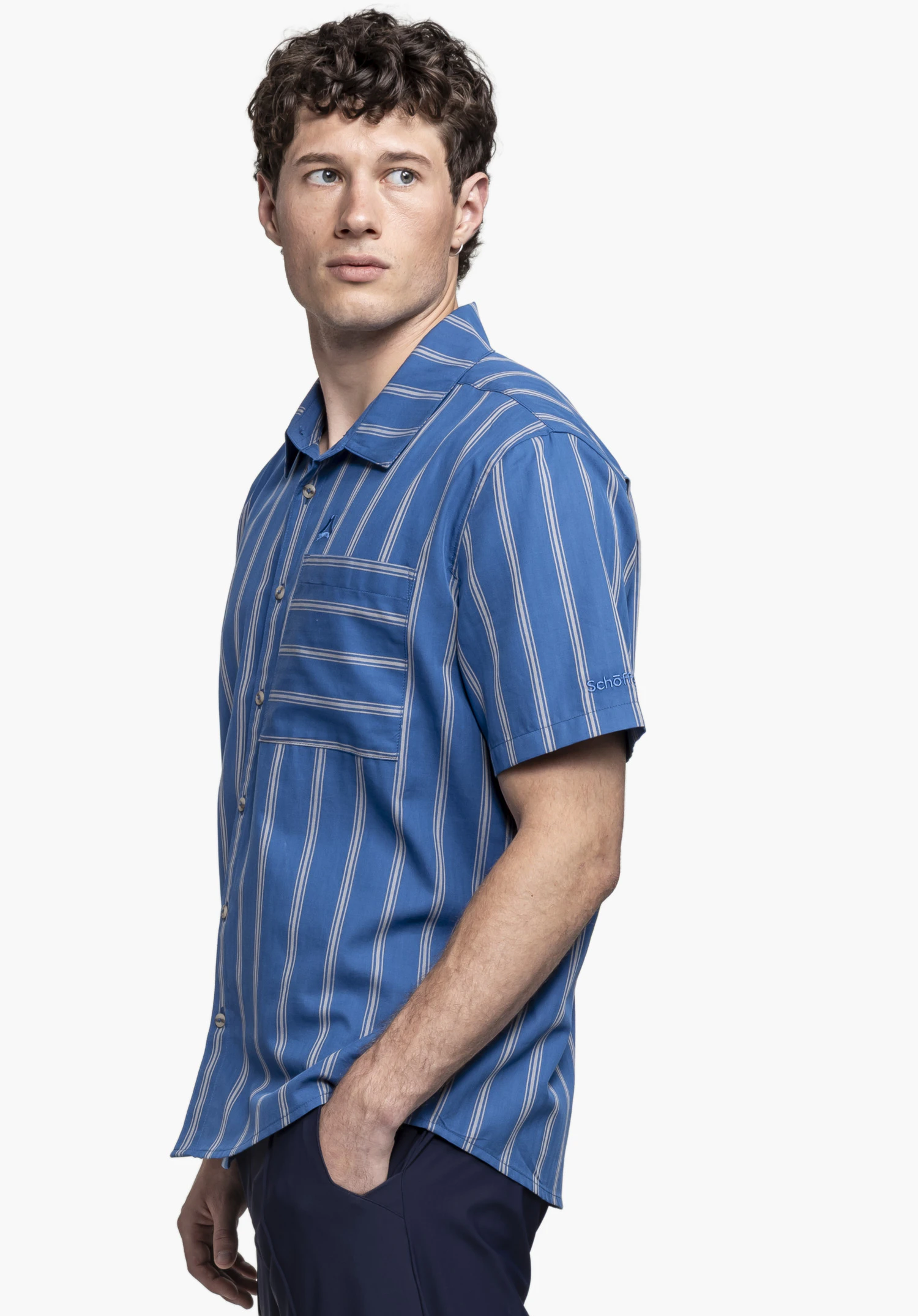Shirt Style Kalmen MEN: Das Model steht seitlich vor einem neutralen Hintergrund und präsentiert das Hemd in Blau mit klassischem Streifenmuster. Das dezente Schöffel-Logo ist gut sichtbar am Ärmel platziert.