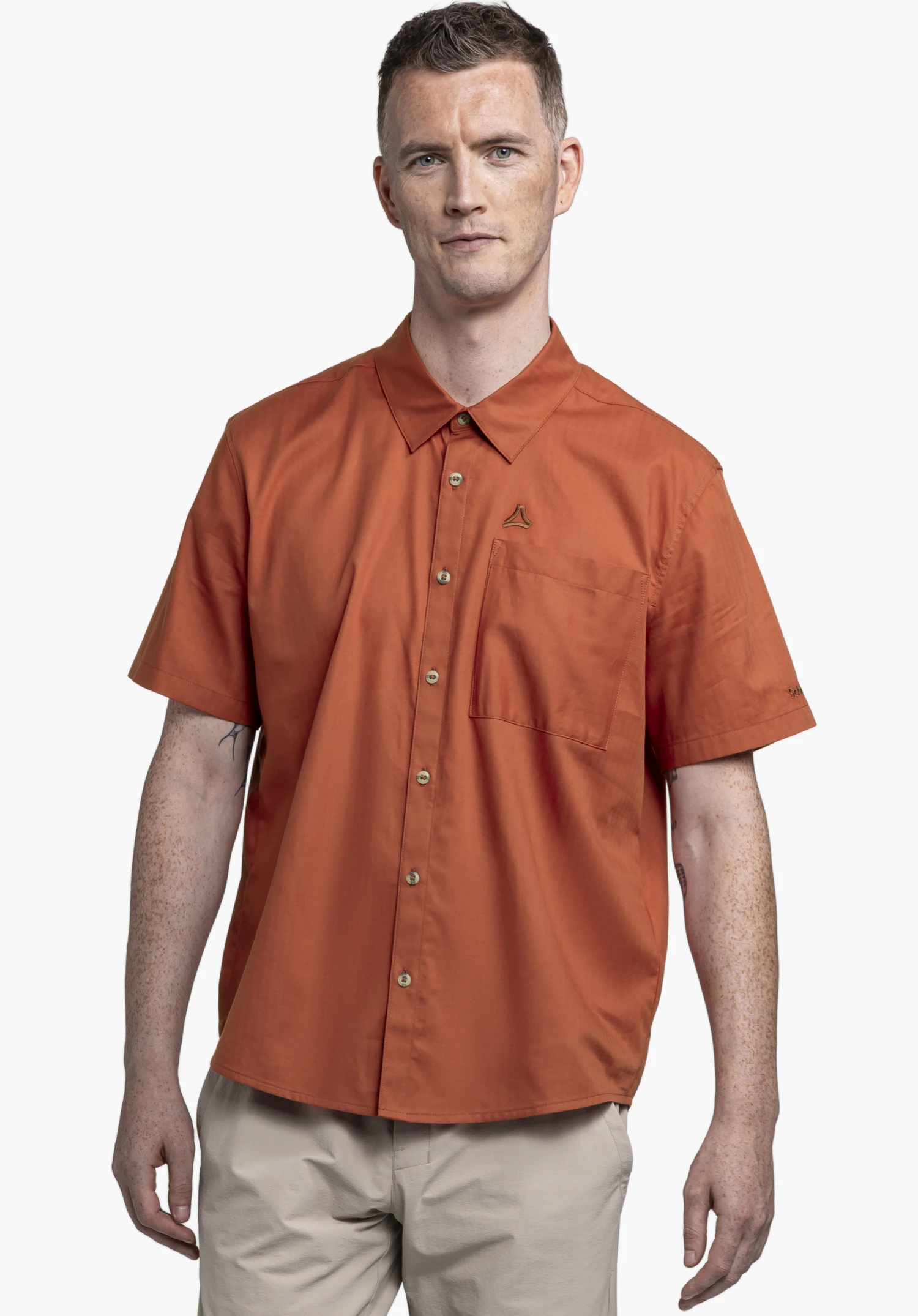 Shirt Style Kalmen MEN : Un mannequin pose de face sur un fond neutre et porte la chemise à manches courtes dans un coloris rouge chaleureux. Le logo Schöffel est apposé avec discrétion sous forme d'icône sur la poche poitrine.