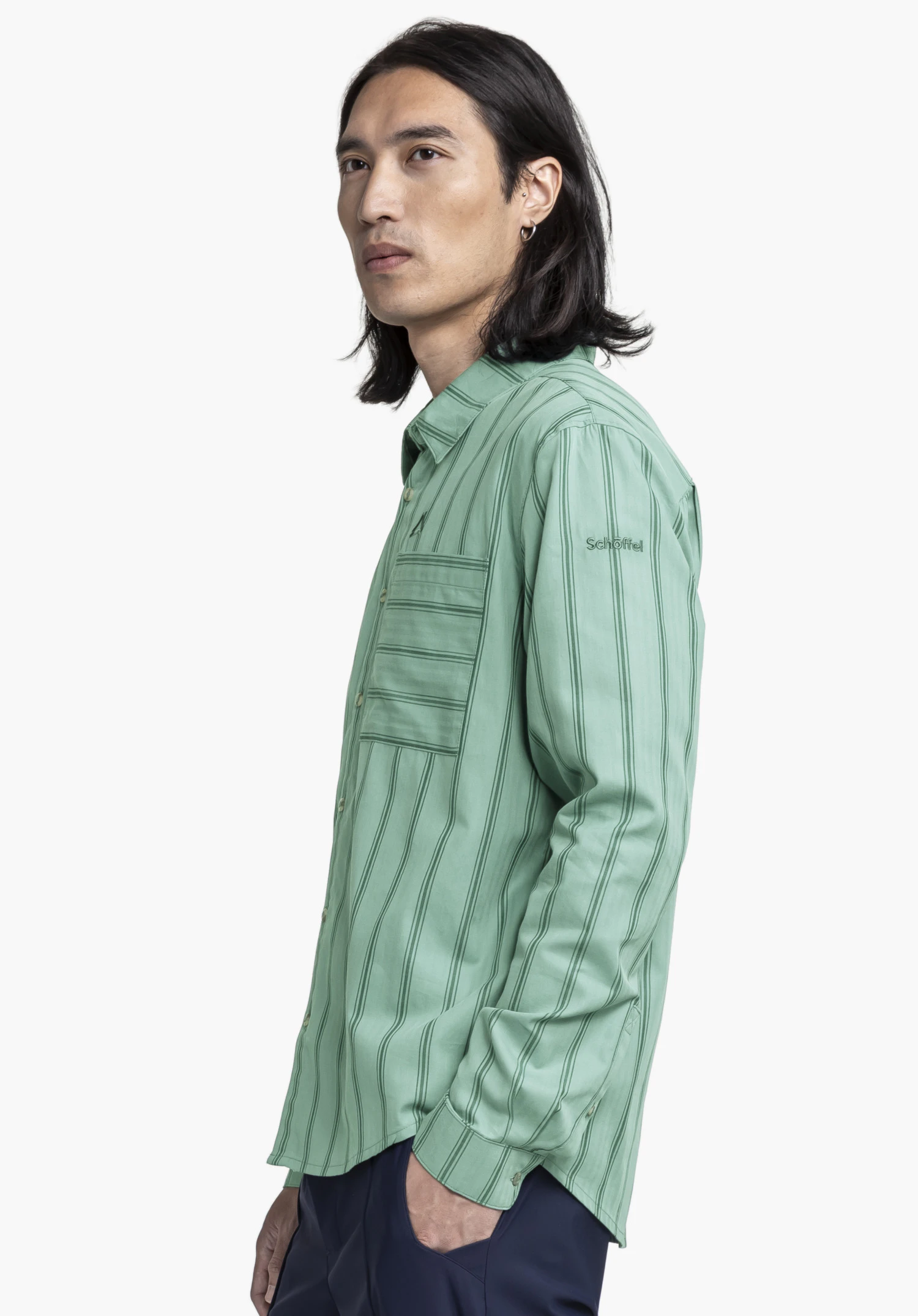 Shirt Style Chanduy MEN en vert. Le modèle pose de profil sur un fond neutre et porte cette chemise à manches longues au motif rayé discret. Le logo Schöffel est brodé sur le haut du bras dans un ton assorti et reste parfaitement visible.