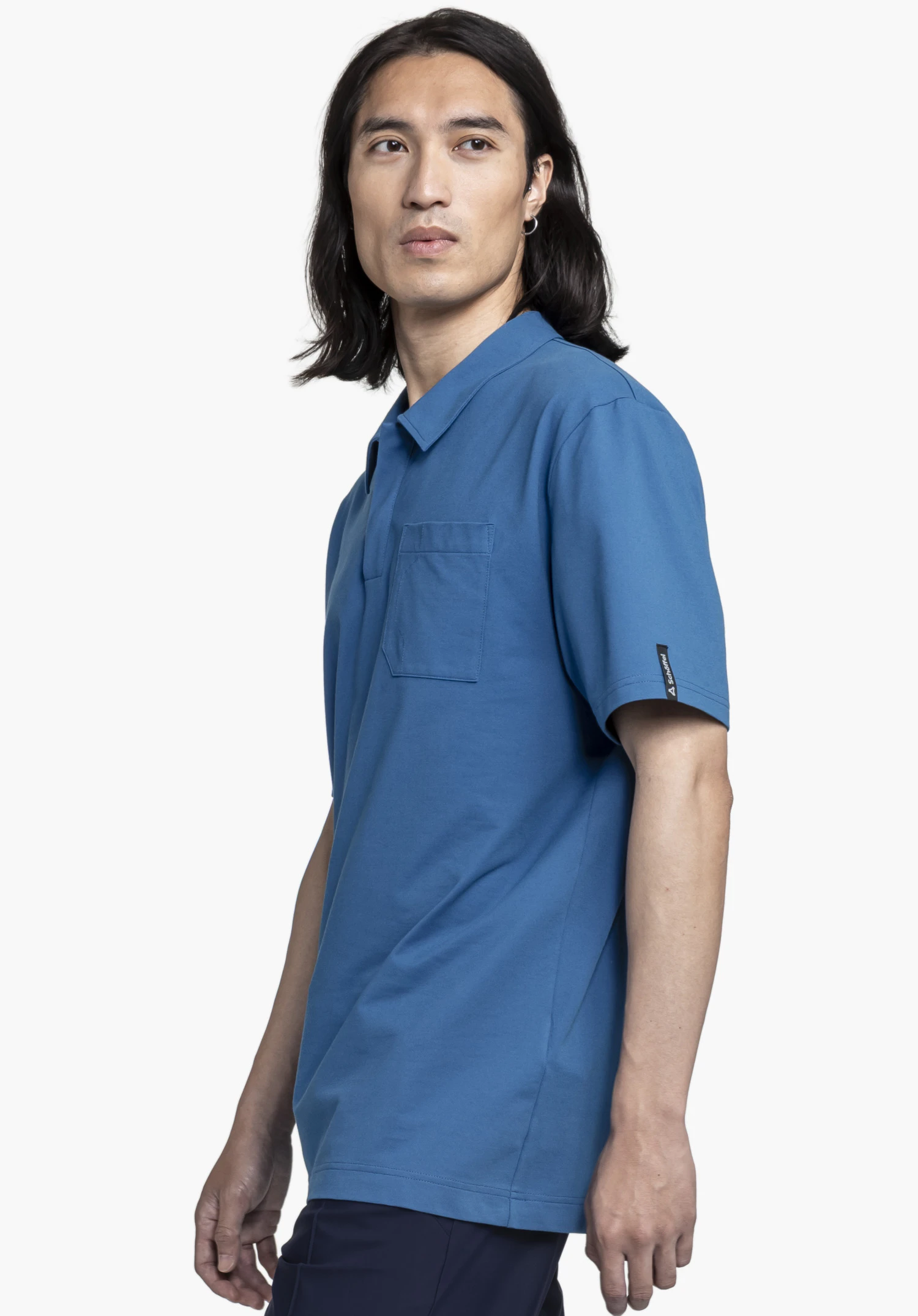 Polo Shirt Style Serenio MEN. A model stands sideways presenting the blue polo shirt with a chest pocket. A small Schöffel logo tag is visible on the left sleeve. The image is set against a neutral background.