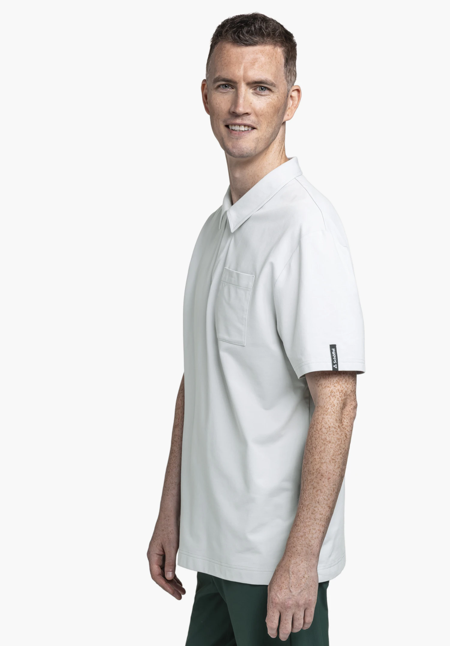 Polo Shirt Style Serenio MEN in grey, worn by a model standing sideways and smiling at the camera. A chest pocket and a small Schöffel-Logo on the left sleeve are visible, presented against a neutral background.