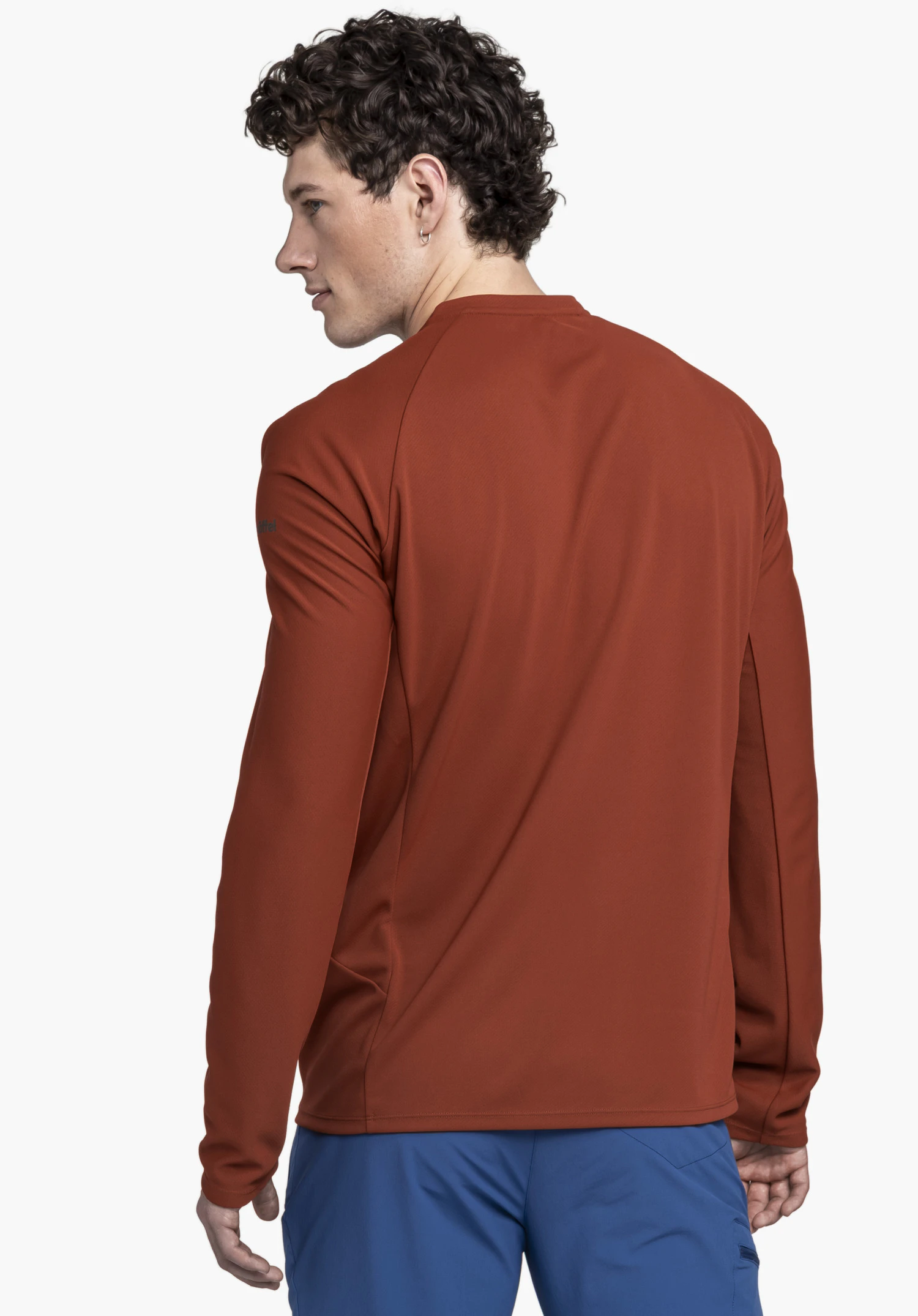 Longsleeve Style Enixa MEN : Un mannequin aux cheveux bouclés porte le haut à manches longues de couleur rouge, vu de dos avec la tête légèrement tournée, sur un fond neutre. Le discret logo Schöffel sur la manche souligne le design sportif idéal pour l'hiver.
