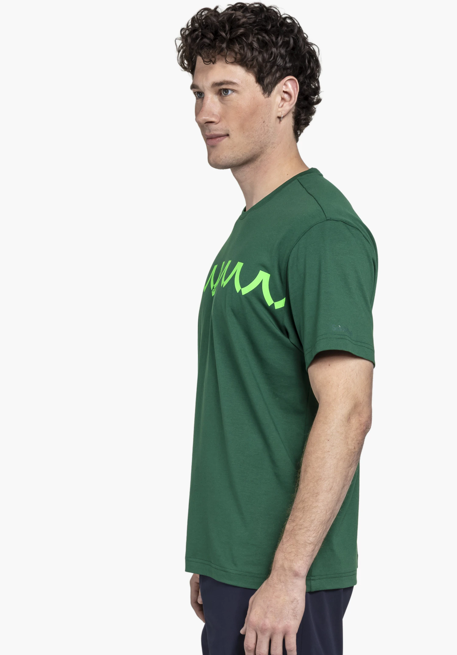 T Shirt Style Elmori MEN en vert présente un homme athlétique photographié de profil sur un fond neutre. Le t-shirt se distingue par son impression frontale marquante, tandis que le logo Schöffel discret sur la manche complète ce look authentique.