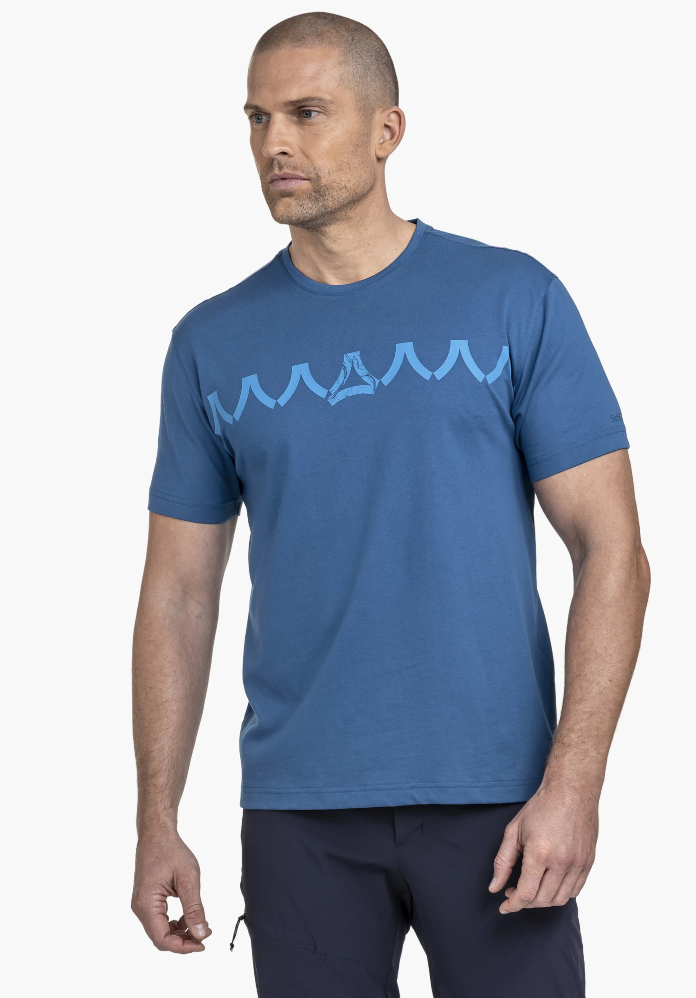 T Shirt Style Elmori MEN en bleu sur un fond neutre. Le mannequin pose légèrement de côté et regarde vers l'avant. Ce t-shirt se distingue par son imprimé moderne sur la poitrine et le logo Schöffel discrètement placé sur la manche.