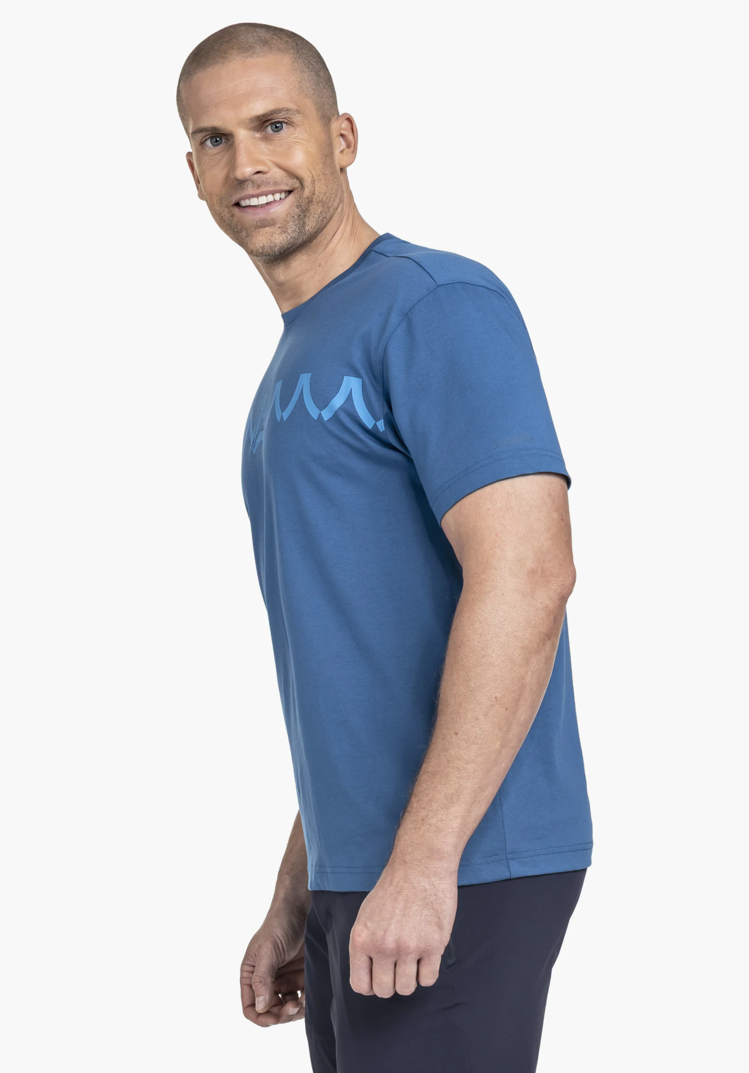 T Shirt Style Elmori MEN en bleu : Le mannequin pose de trois-quarts face devant un fond neutre, mettant en valeur le t-shirt avec un logo Schöffel distinctif sur la poitrine. La coupe décontractée souligne le look sportif.