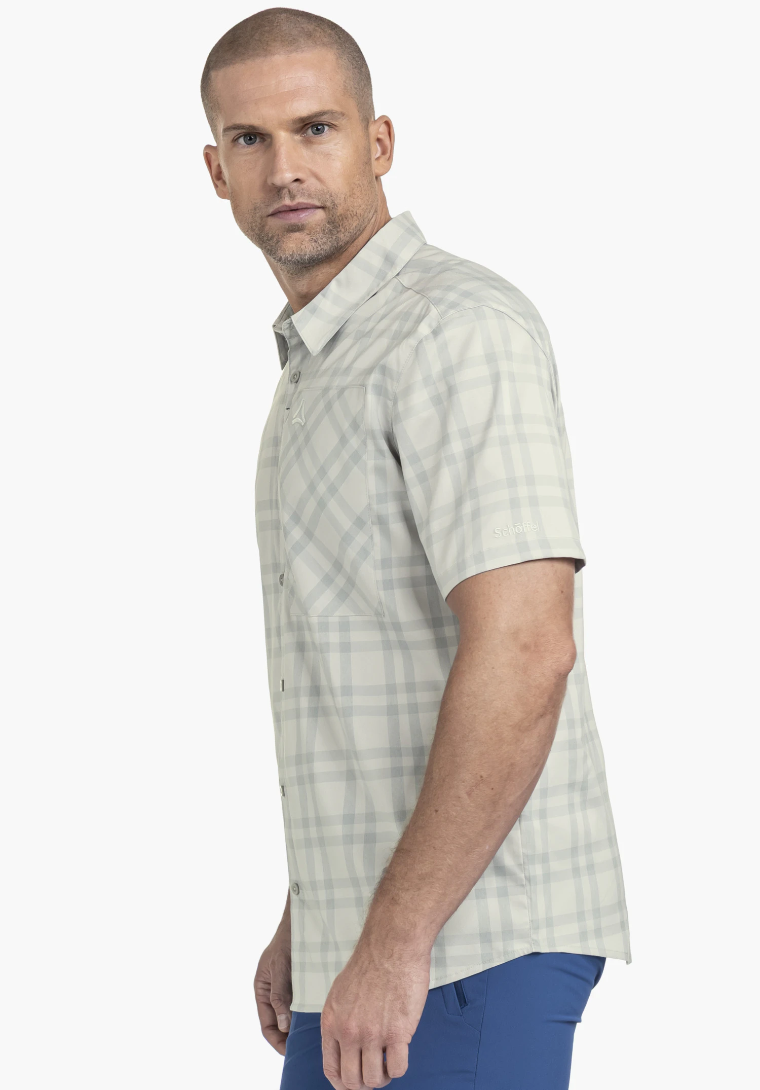 Shirt Style Frolya MEN, porté par un mannequin qui se tient de profil et regarde vers l'appareil photo. La chemise à manches courtes à carreaux en gris présente le logo Schöffel sur la poitrine et la manche, présentée sur un fond neutre.