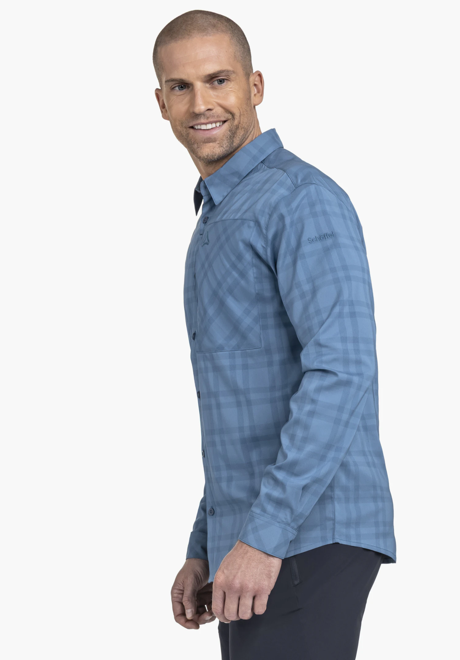 Shirt Style Fraris MEN worn by a model standing sideways and smiling at the camera. The shirt in blue features a subtle check pattern and displays the Schöffel logo on the left upper arm. The shot is taken against a neutral background.