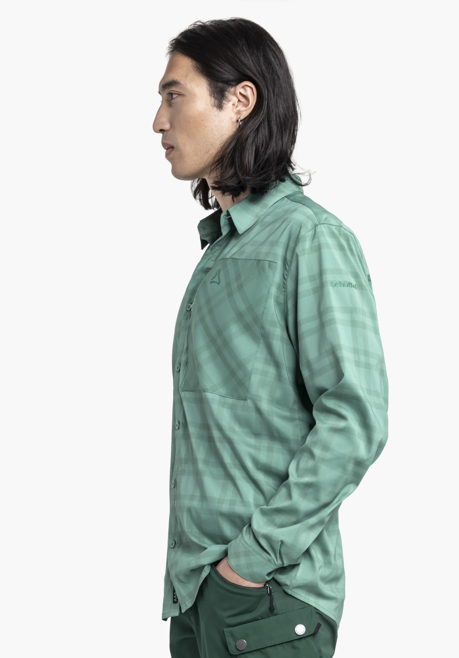 Shirt Style Fraris MEN: Das Modell steht seitlich vor einem neutralen Hintergrund und präsentiert das Shirt in elegantem Grün mit dezentem Karomuster. Das hochwertige Material und das diskret aufgestickte Schöffel-Logo am Ärmel unterstreichen den funktionalen Stil.