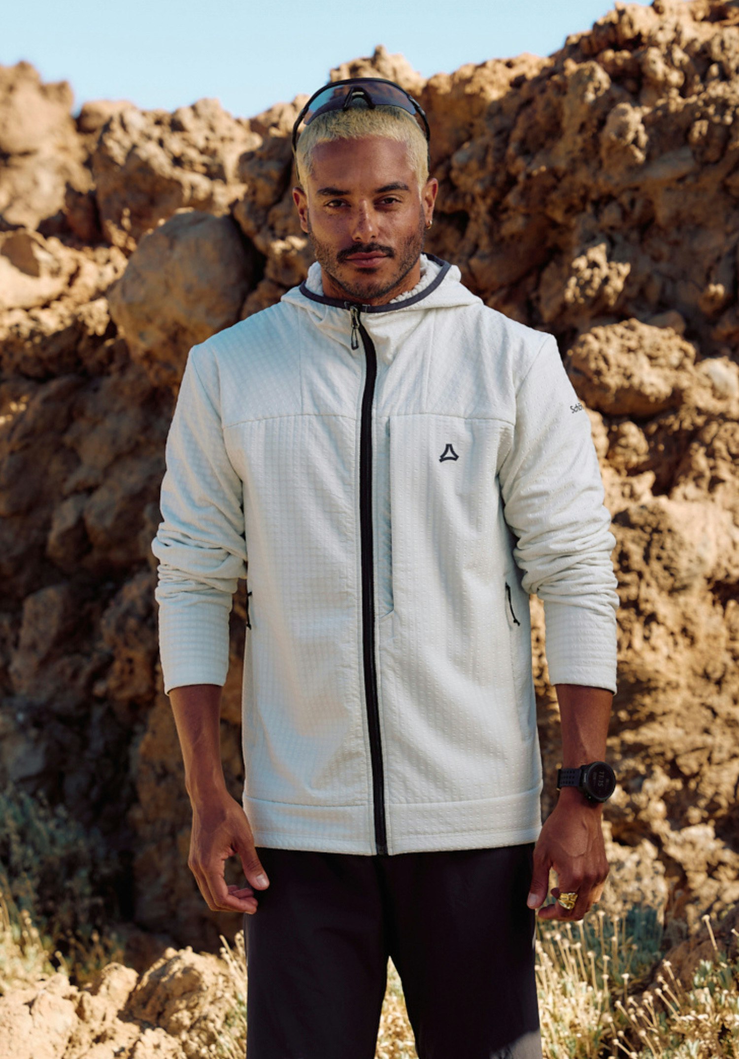 Fleece Hoody Style Enixa MEN. Un mannequin se tient de face et présente le sweat à capuche texturé dans la couleur natur avec une fermeture éclair frontale noire. Le Schöffel-Logo noir est bien visible sur la poitrine et la manche gauche. Le mannequin regarde légèrement sur le côté et se tient devant un fond neutre.